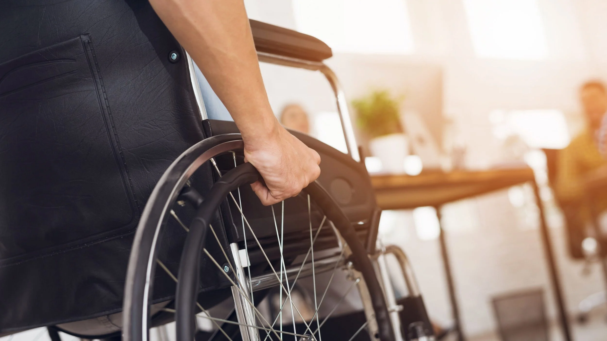 Close-up of a person's hand gripping the wheel of a wheelchair, with a blurred indoor office background.