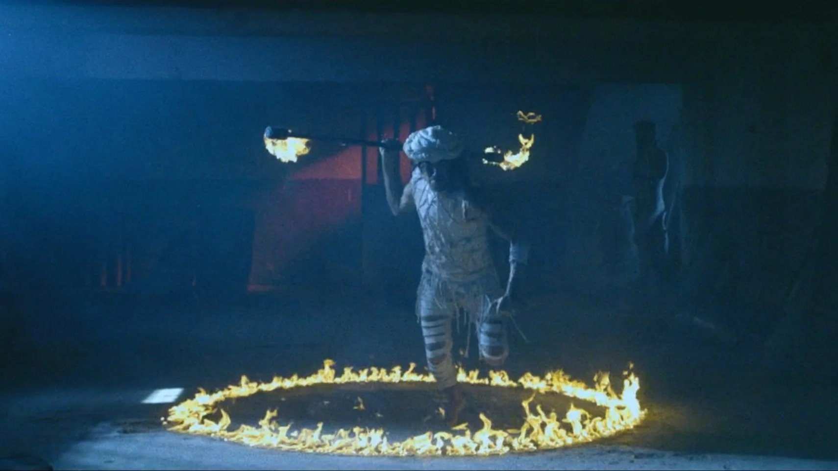 Persona disfrazada de payaso con ropa rota dentro de un círculo de fuego en un entorno oscuro.