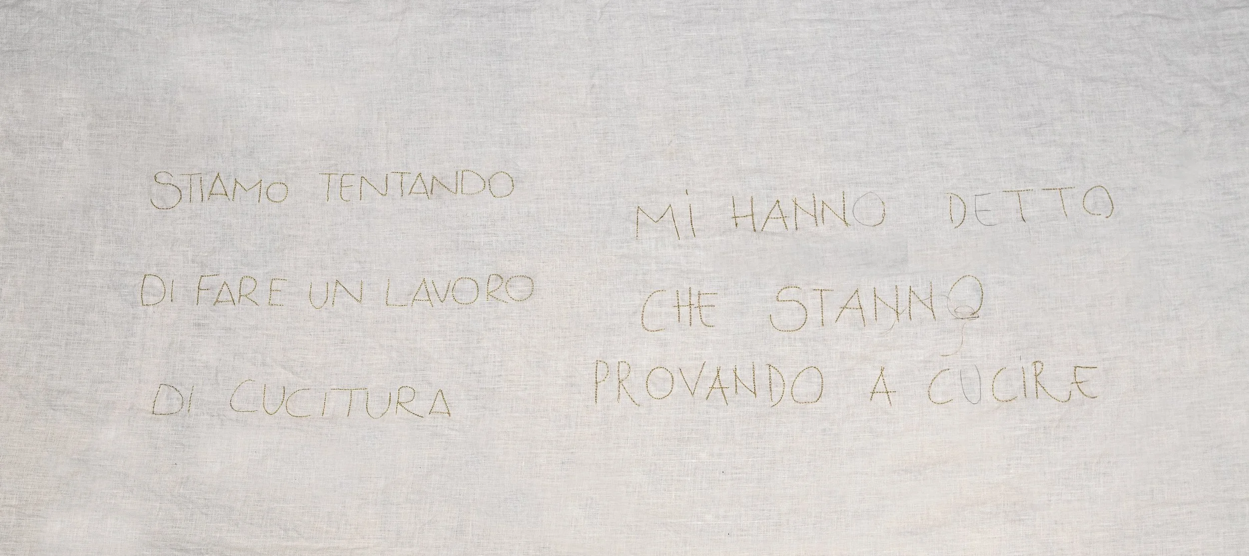 tessuto bianco con scritte ricamate in italiano; il testo dice 'stiamo tentando di fare un lavoro di cucitura mi hanno detto che stanno provando a cucire'