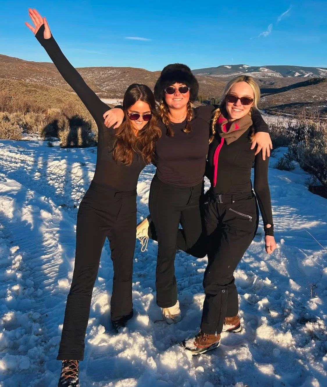 Three women smiling and embracing in a snowy outdoor landscape with hills in the background, wearing winter clothing and sunglasses.