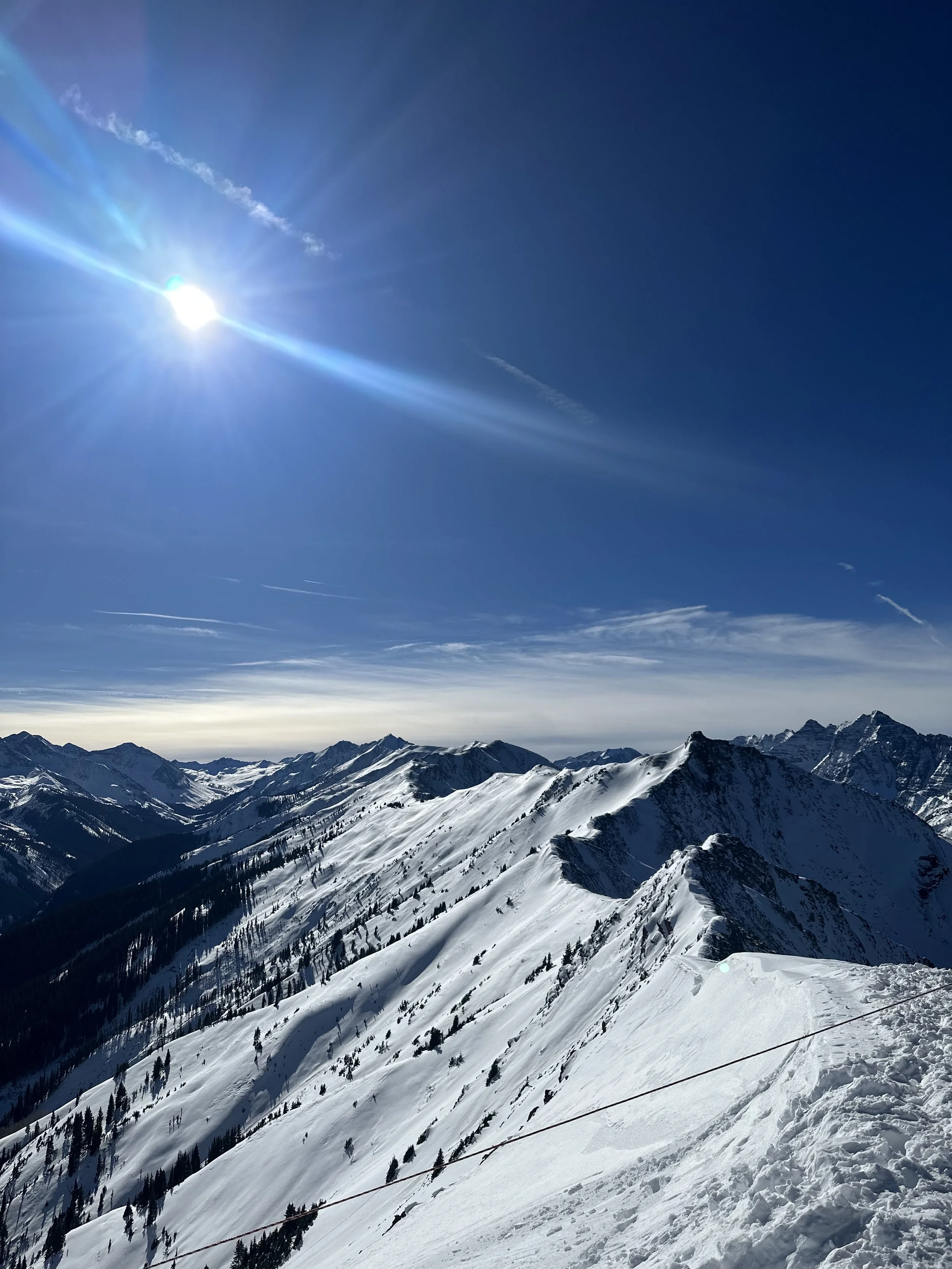 Snow-covered mountain range under a bright, sunny sky with visible sun rays and some contrails.