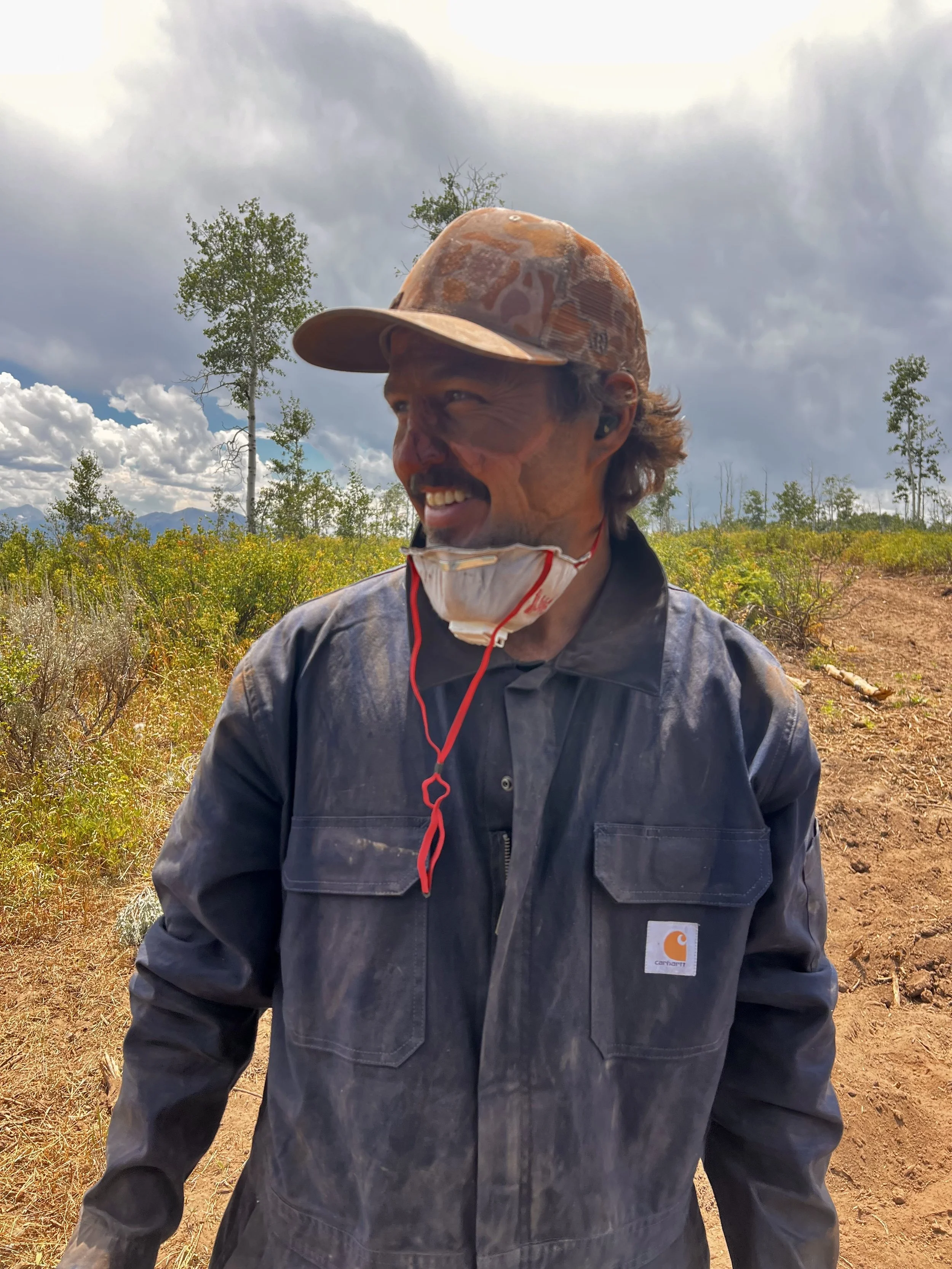Smiling man with outdoor field, trees, and cloudy sky in the background, wearing a camouflage cap, gray Carhartt jacket, and a face mask hanging around his neck.