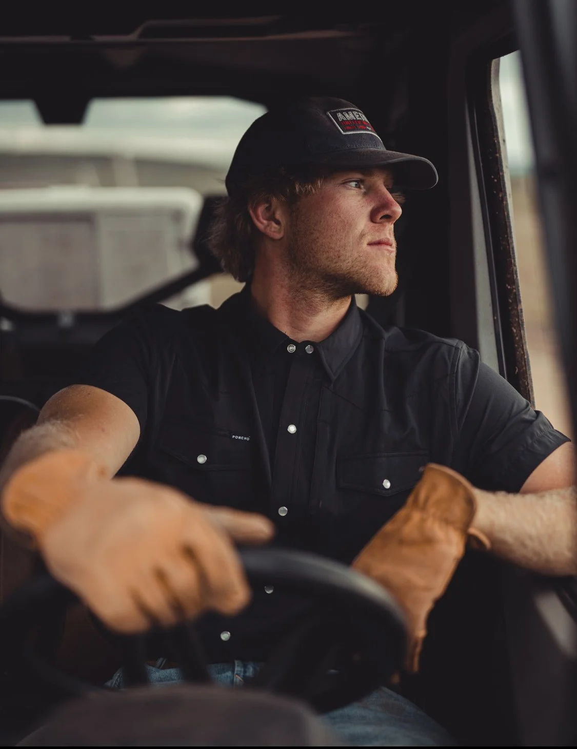 A man wearing a black cap and black short-sleeve shirt driving a vehicle with brown gloves on his hands, looking out the window.