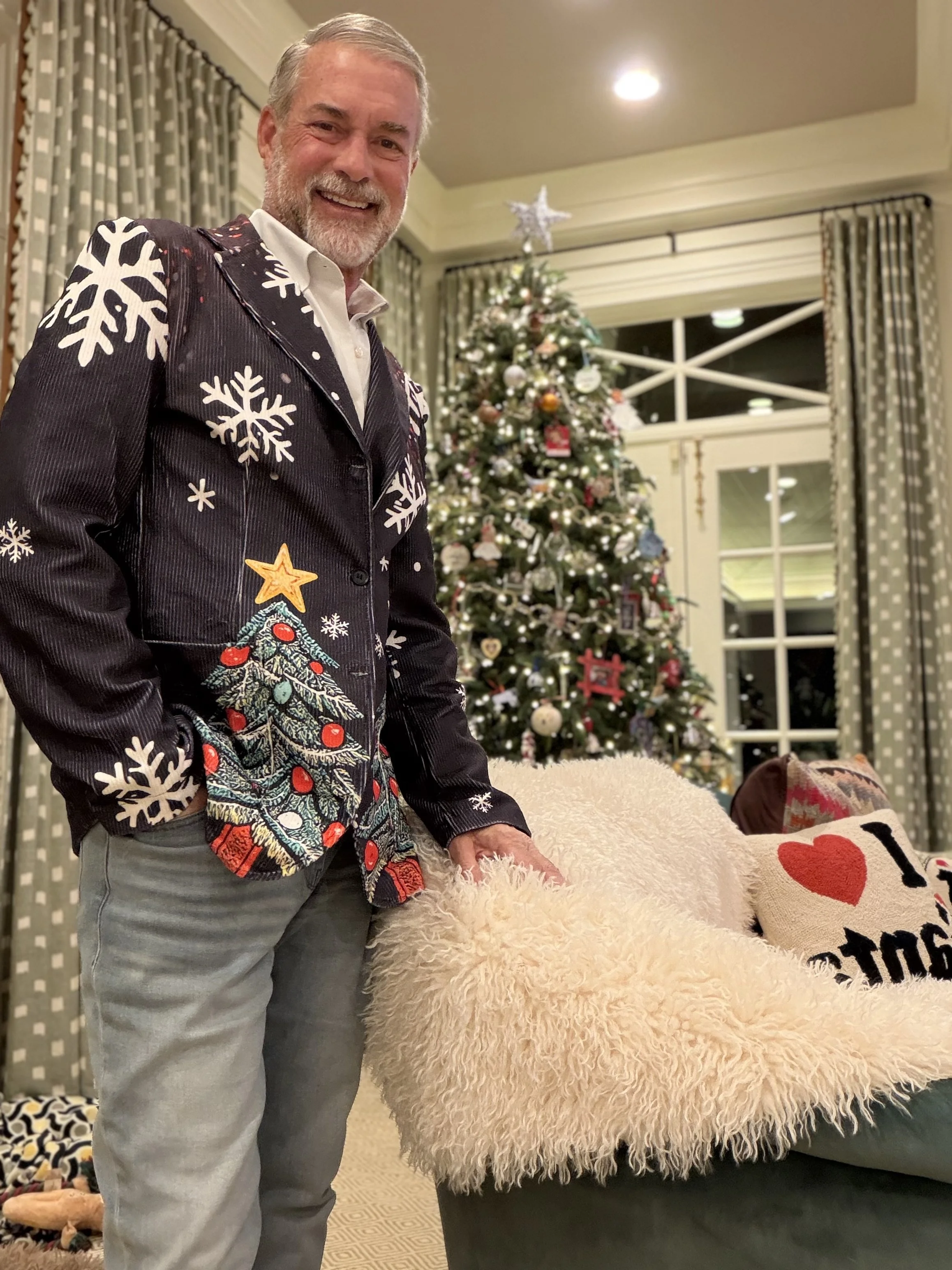 A man with gray hair and beard smiling, wearing a black Christmas sweater with Christmas tree and snowflake patterns, standing in a decorated living room with a Christmas tree in the background, and a beige fluffy blanket and decorative pillow on a s