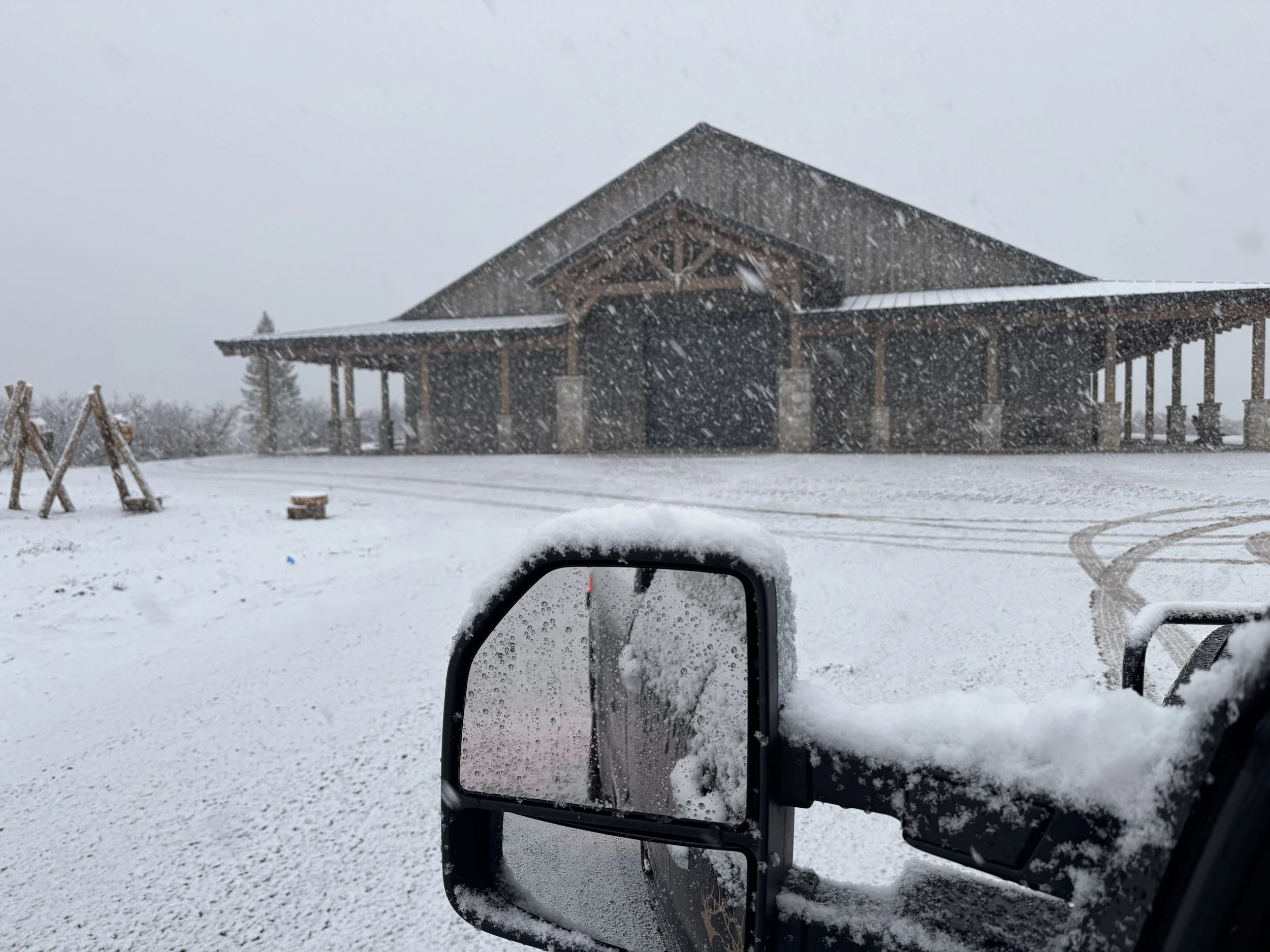 A snow-covered landscape showing a wooden building in the distance with snow falling. In the foreground, a snow-covered vehicle side mirror is visible.