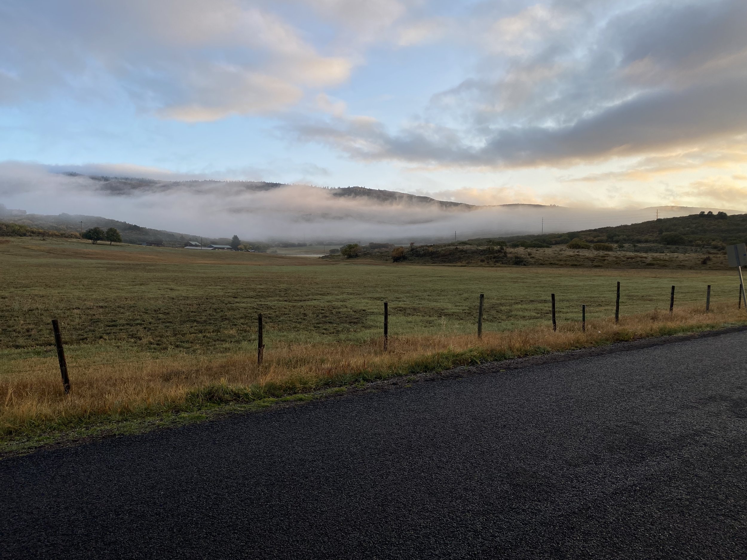 Open field with a fence, misty hills in the background, and a cloudy sky at sunrise or sunset.