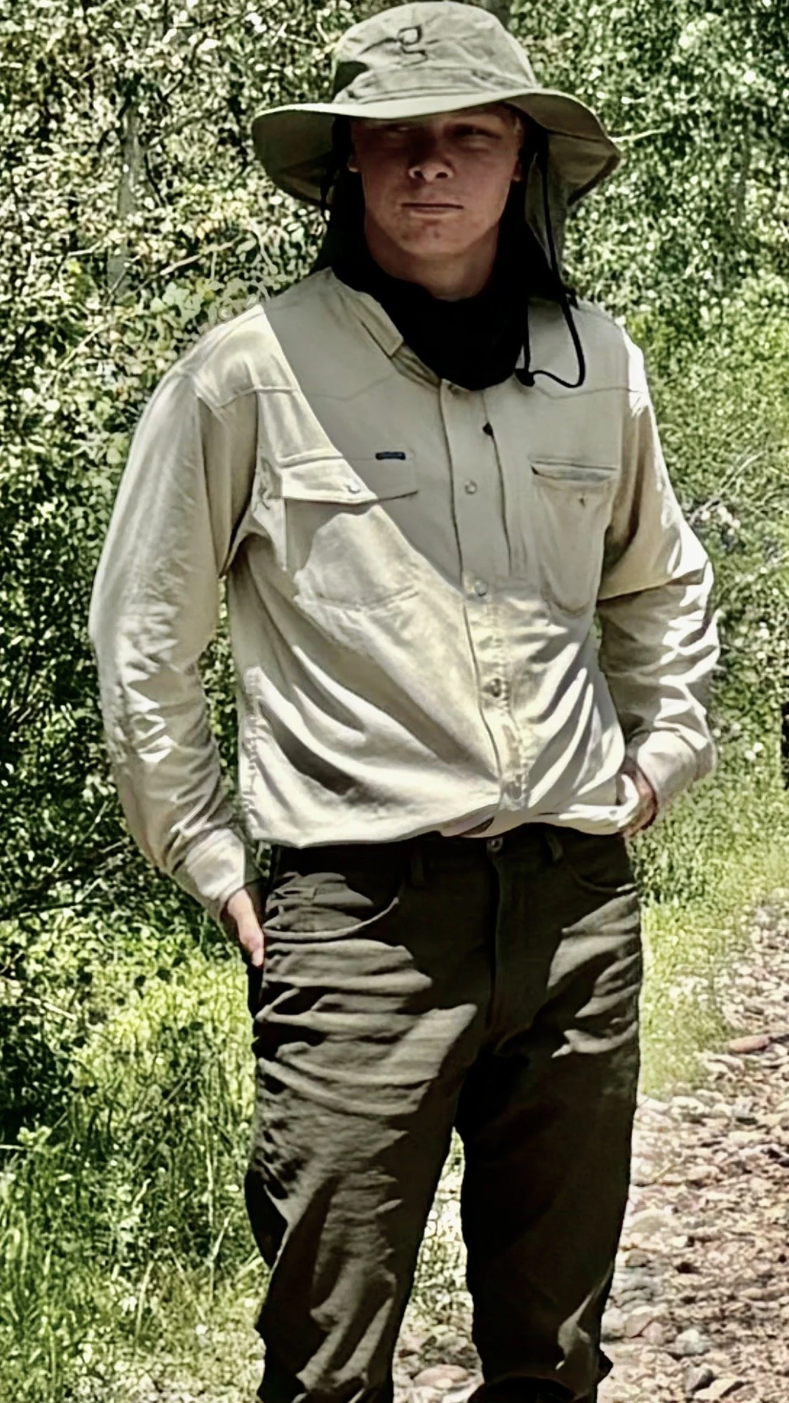 Person wearing a wide-brimmed hat, beige long-sleeve shirt, and dark pants standing outdoors on a rocky path surrounded by greenery.