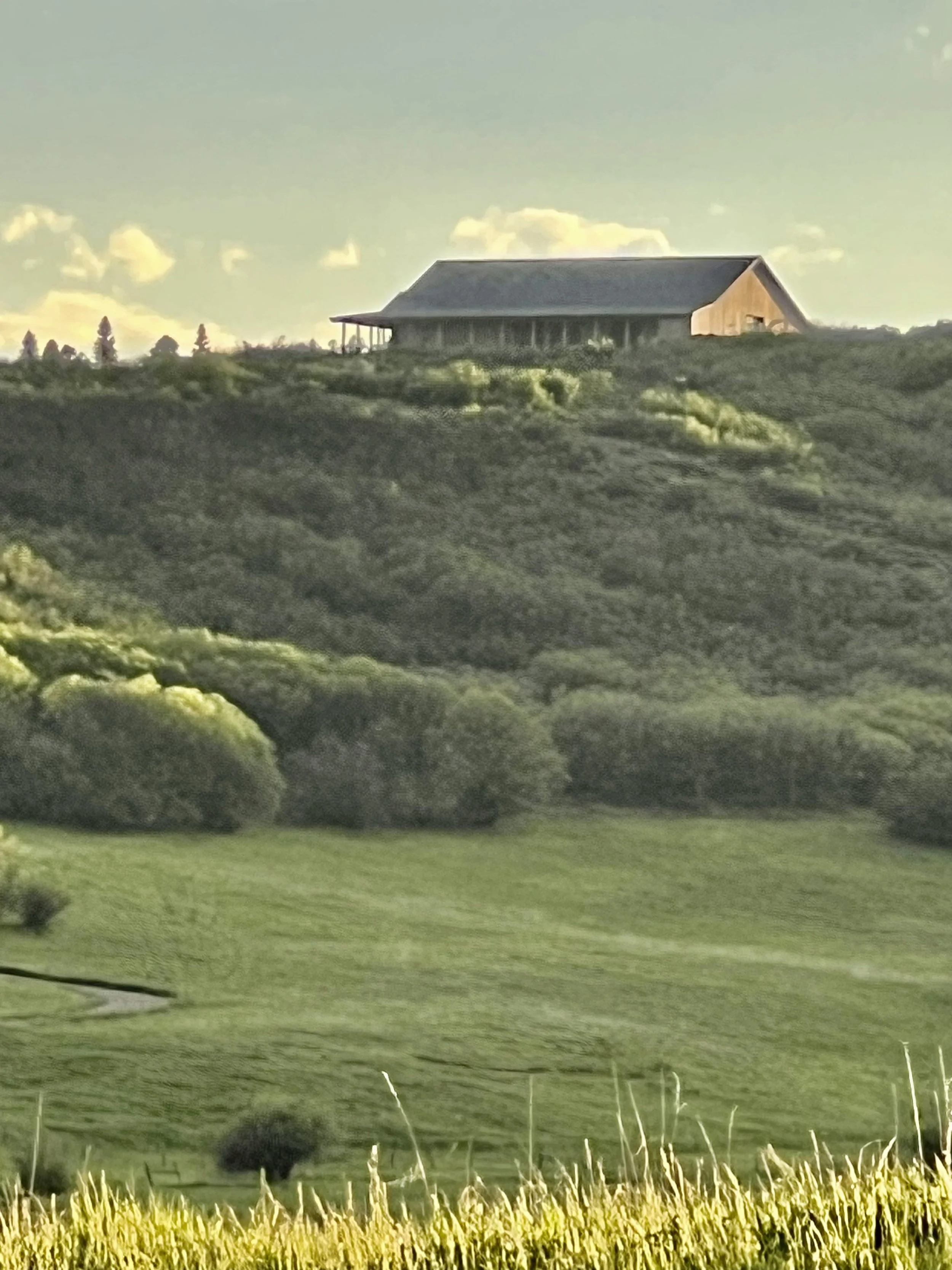 A rural landscape with green rolling hills, trees, and a large barn on the top of the hill under a partly cloudy sky.