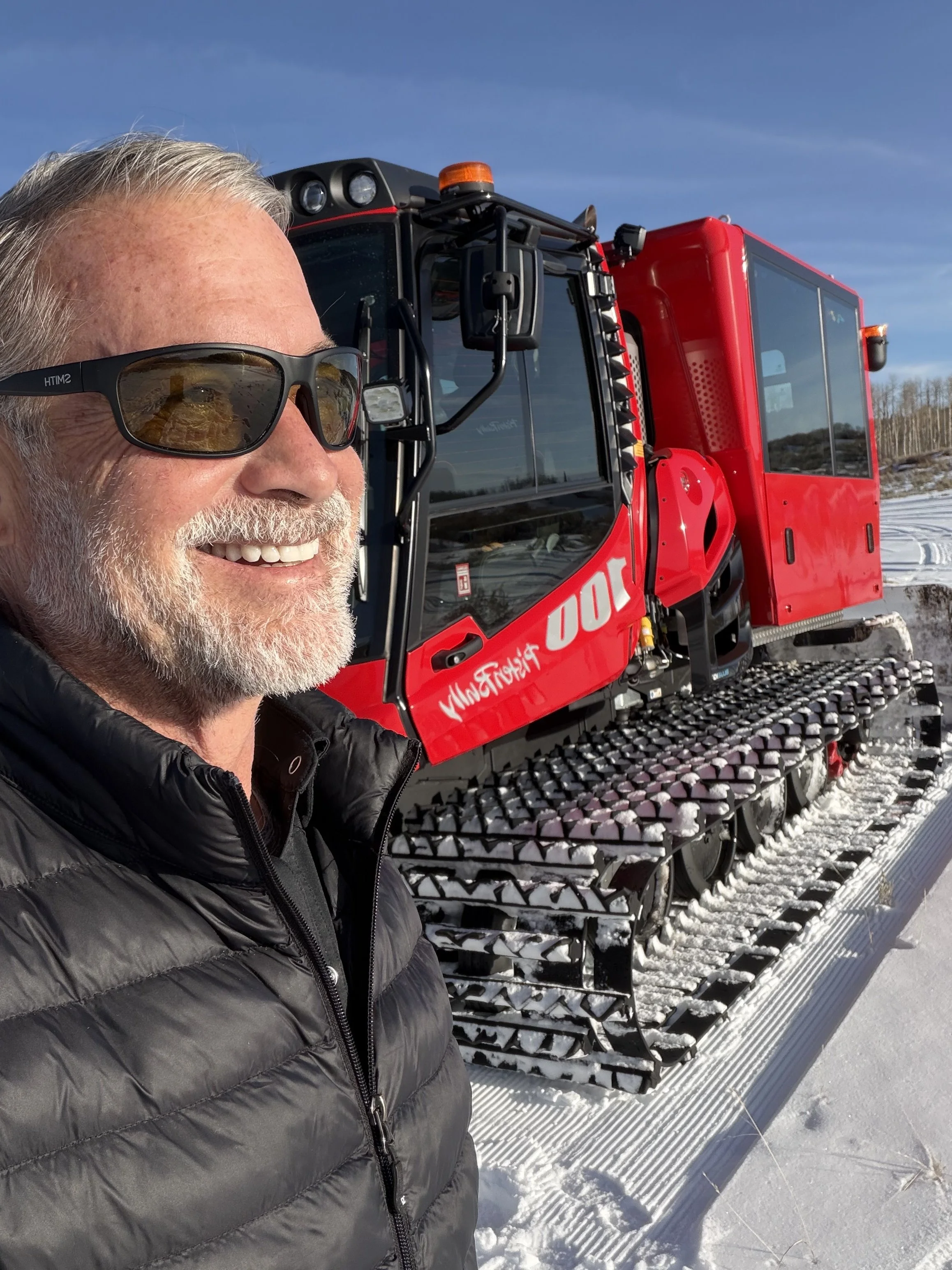 Smiling man with sunglasses taking selfie in front of a red snow grooming machine on a snowy landscape.