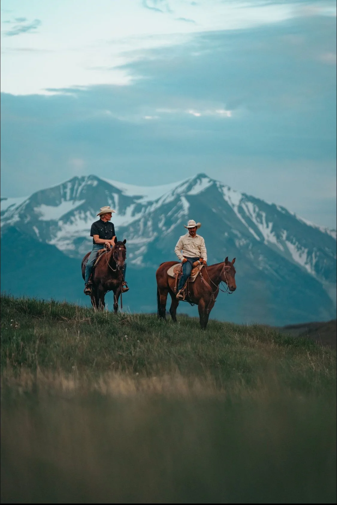 Two men in cowboy hats riding horses on a grassy hill with snow-capped mountains in the background.