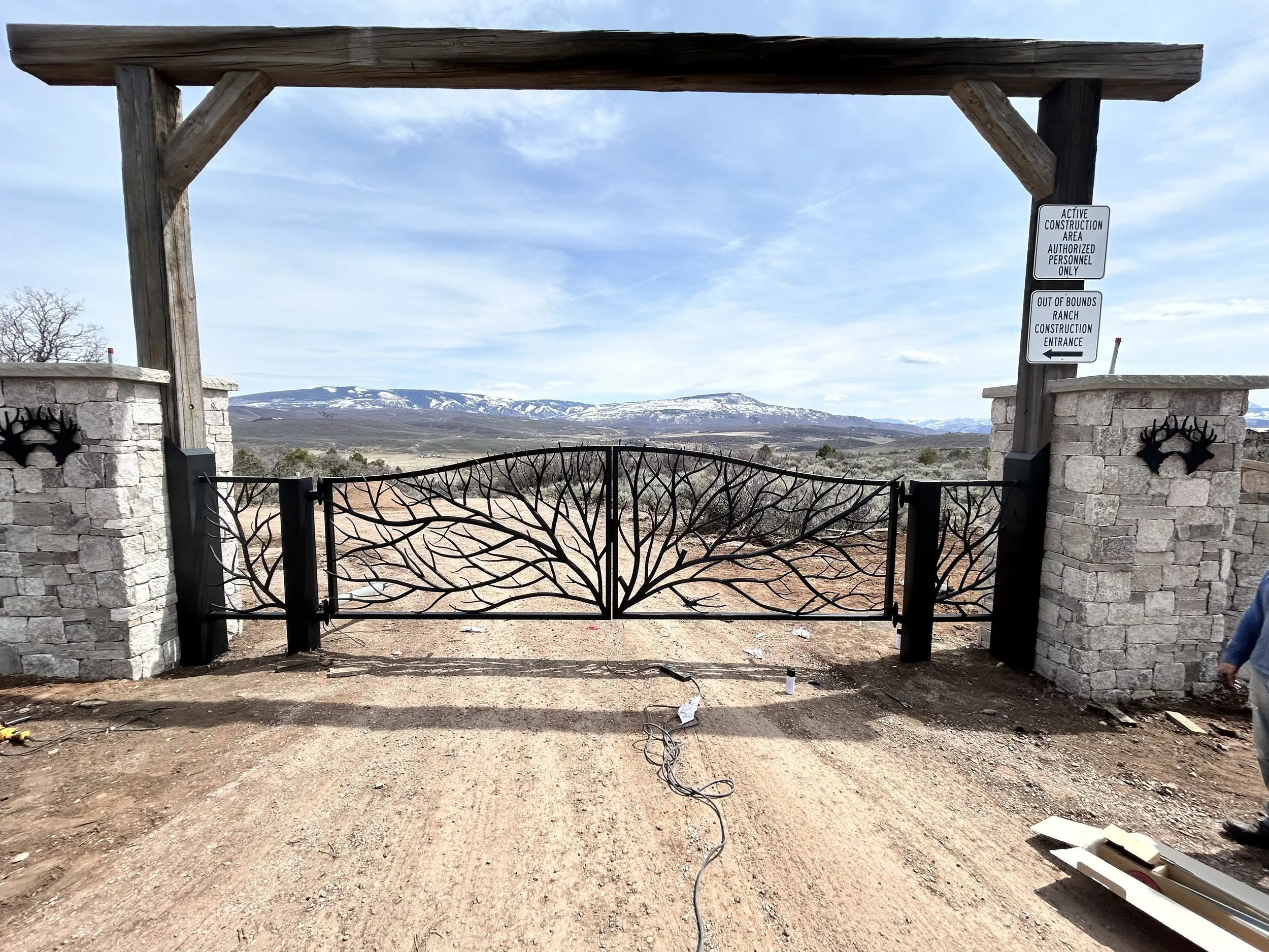 Entrance gate with stone pillars and wooden arch, leading into a rural area with mountains in the distance and snow patches on the peaks.