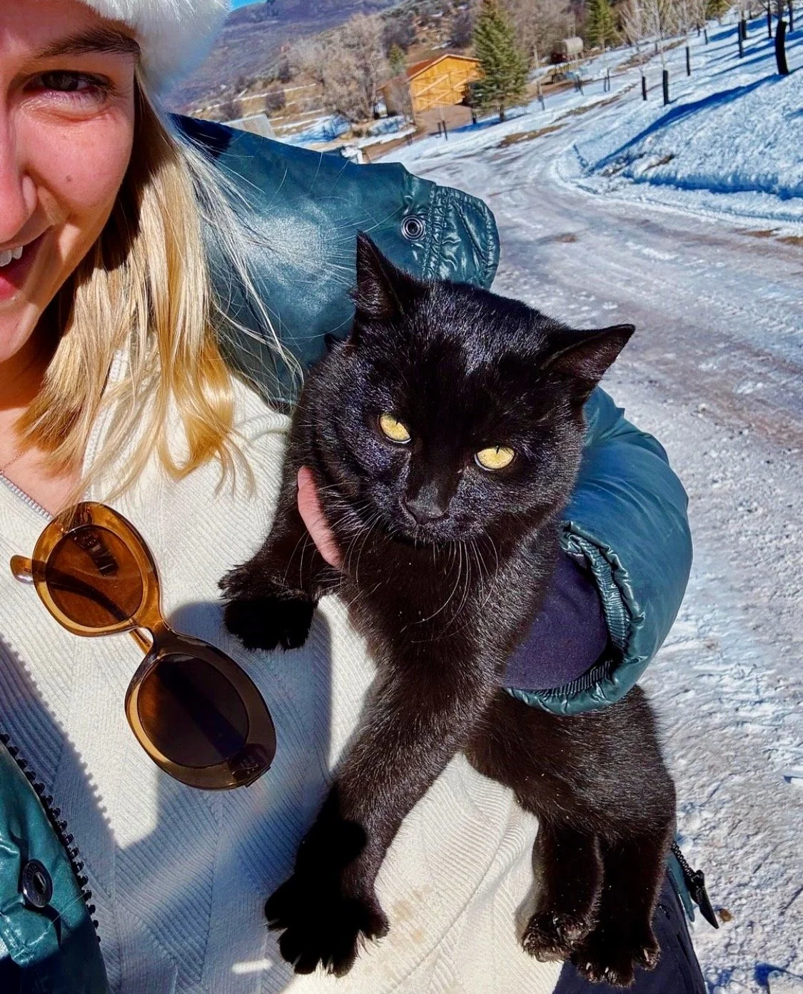 A woman with blonde hair and a white jacket holding a black cat outdoors on a snowy landscape with mountains, trees, and a small building in the background.