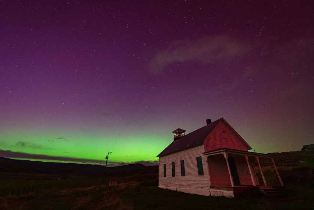 A small house with a porch in a rural landscape at night under a colorful sky illuminated by the Northern Lights, with stars visible overhead.