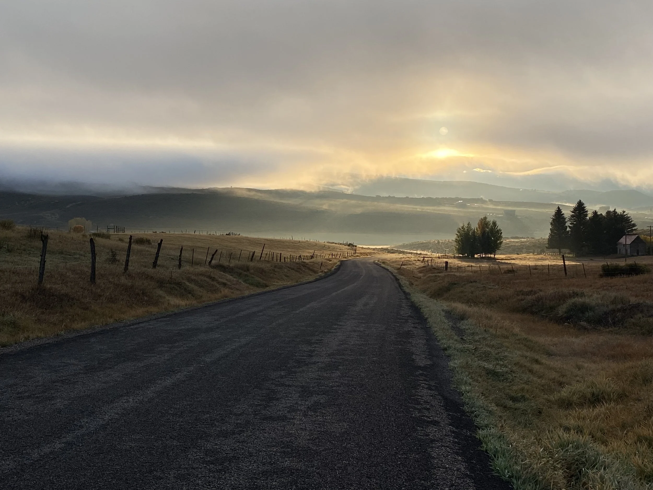 A winding rural road through grassy fields with fences, trees, and distant mountains under a cloudy sky with the sun partially obscured.