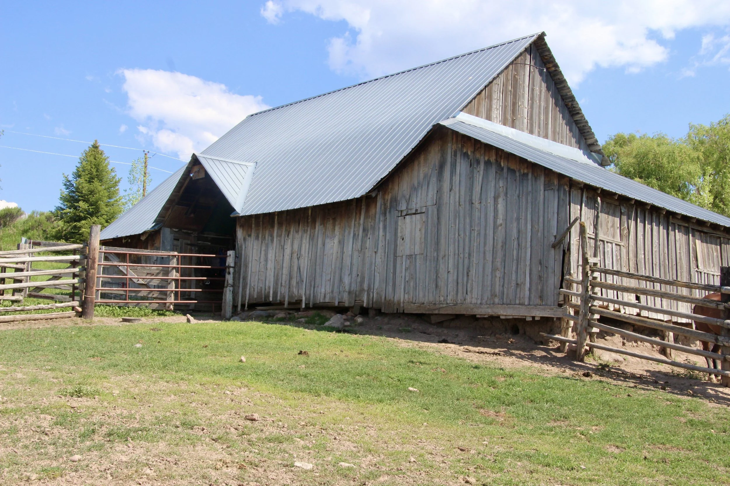 An old wooden barn with a metal roof on a grassy hill under a blue sky with clouds.
