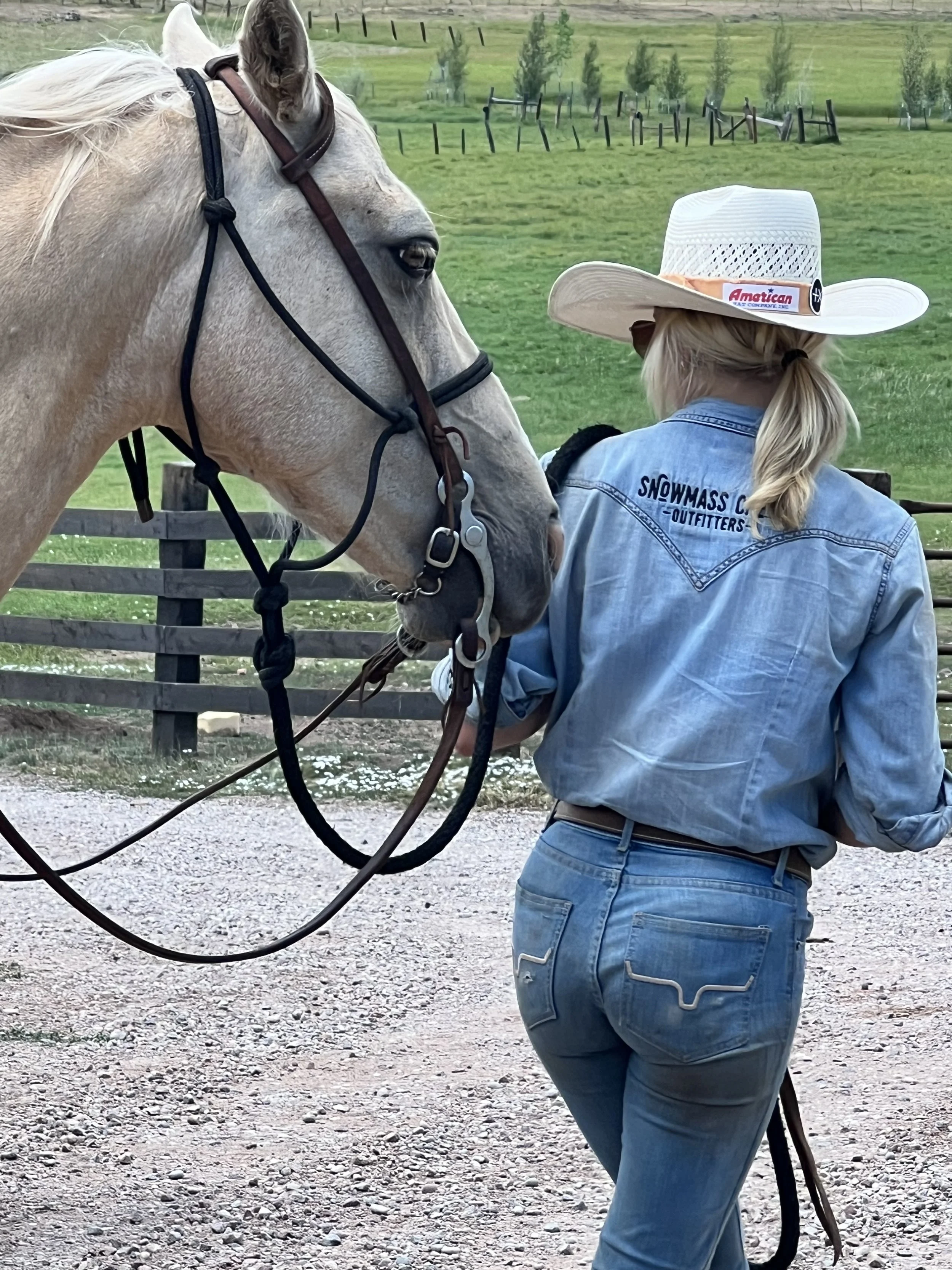 A woman wearing a cowboy hat and a blue denim shirt with 'Snowmass Co Outfitters' on the back stands next to a white horse with a brown mane, holding its reins, in an outdoor rural setting with grassy fields and a wooden fence.