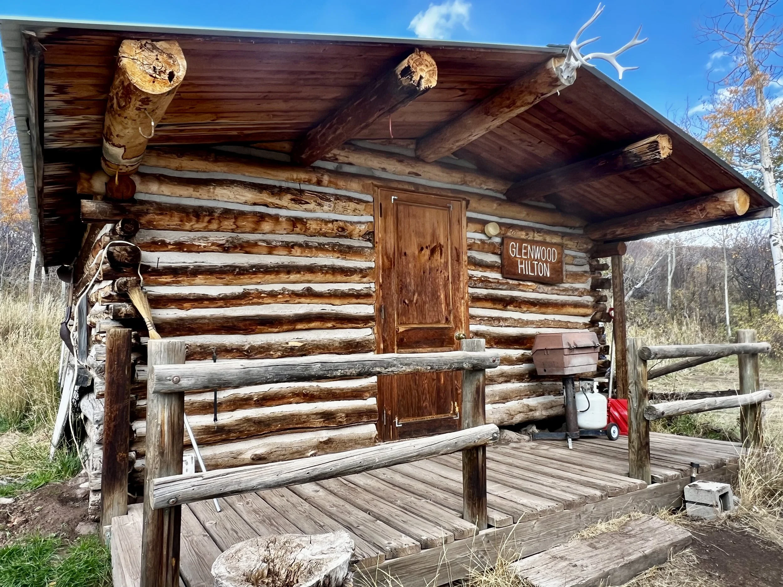 A rustic log cabin with a wooden porch, a sign that reads 'Glenwood Hilton,' and decorative antlers mounted on the roof corner. The cabin is surrounded by trees and grass, with a clear blue sky overhead.