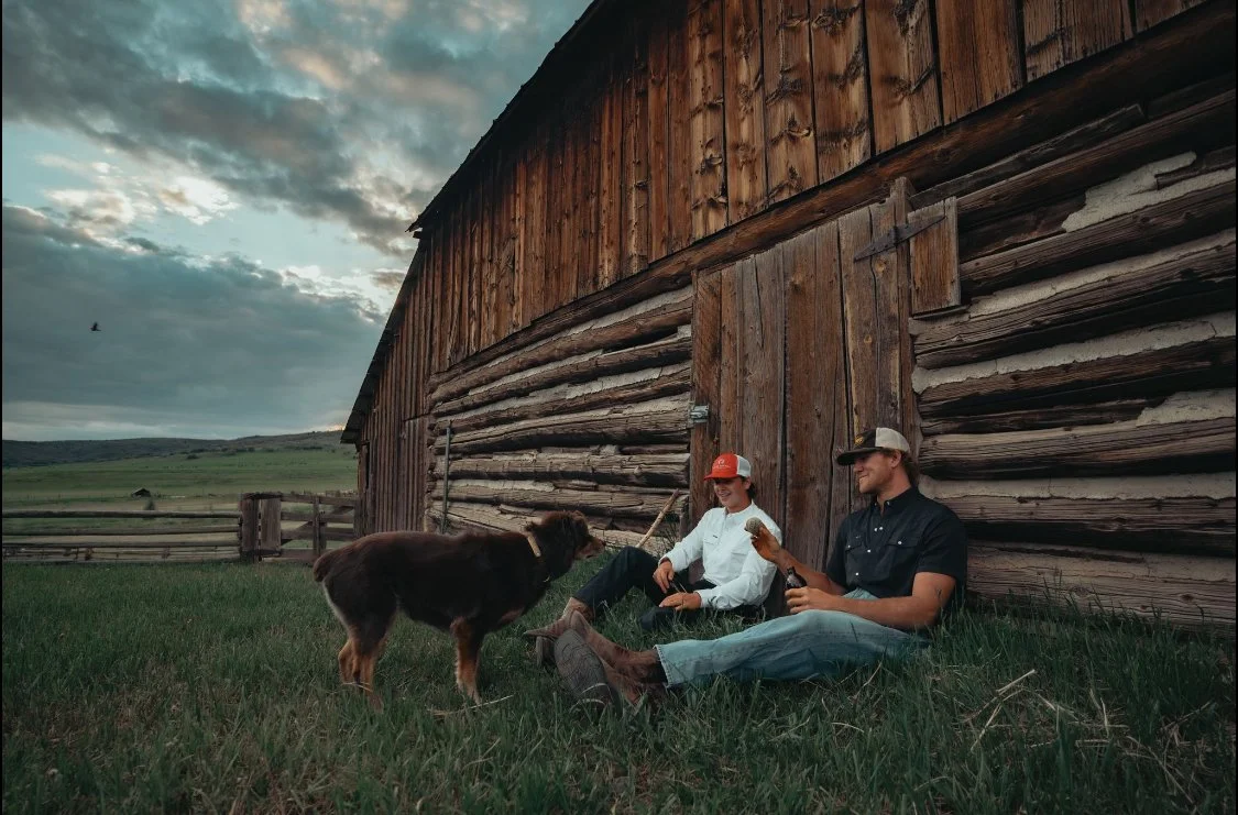 A man and woman sit against a rustic wooden barn, relaxing with a dog while holding drinks. The sky is cloudy, and the scene is rural with open fields in the background.