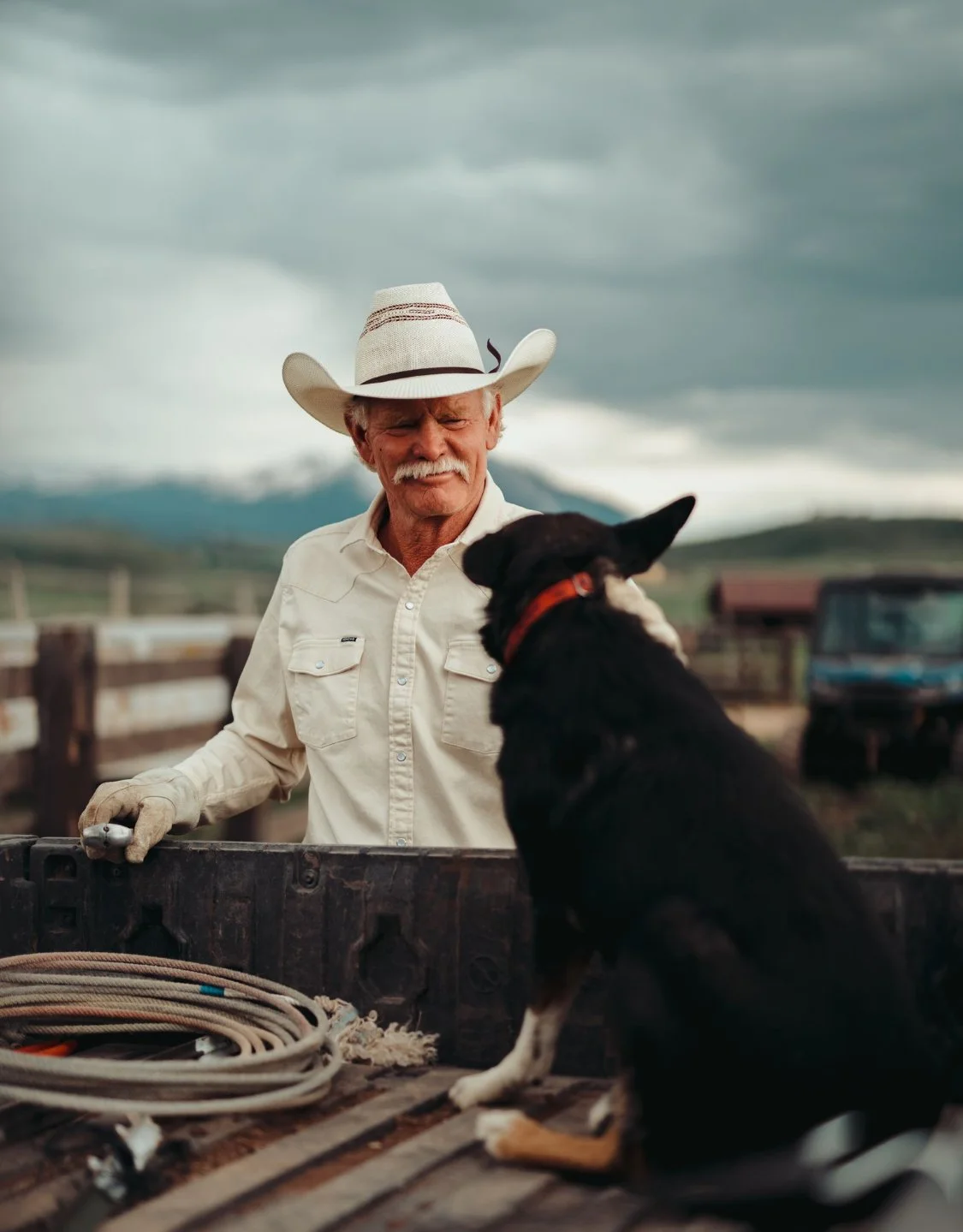 An older man with a white cowboy hat and a light-colored shirt stands by a truck bed, talking to a black dog with a red collar, with a rural landscape and cloudy sky in the background.