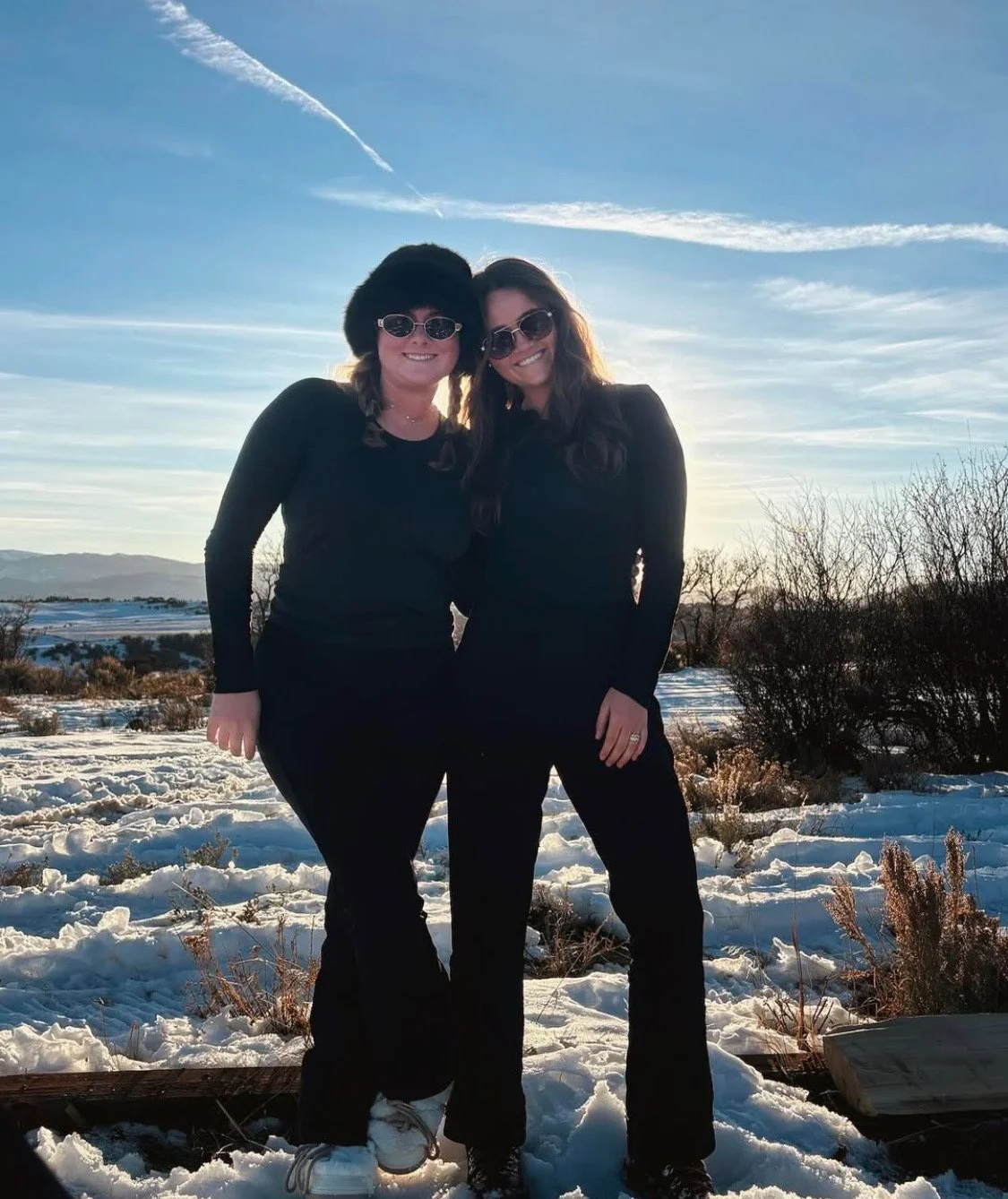 Two women standing outdoors on snowy ground, smiling, wearing black outfits and sunglasses, with a scenic winter landscape and blue sky in the background.