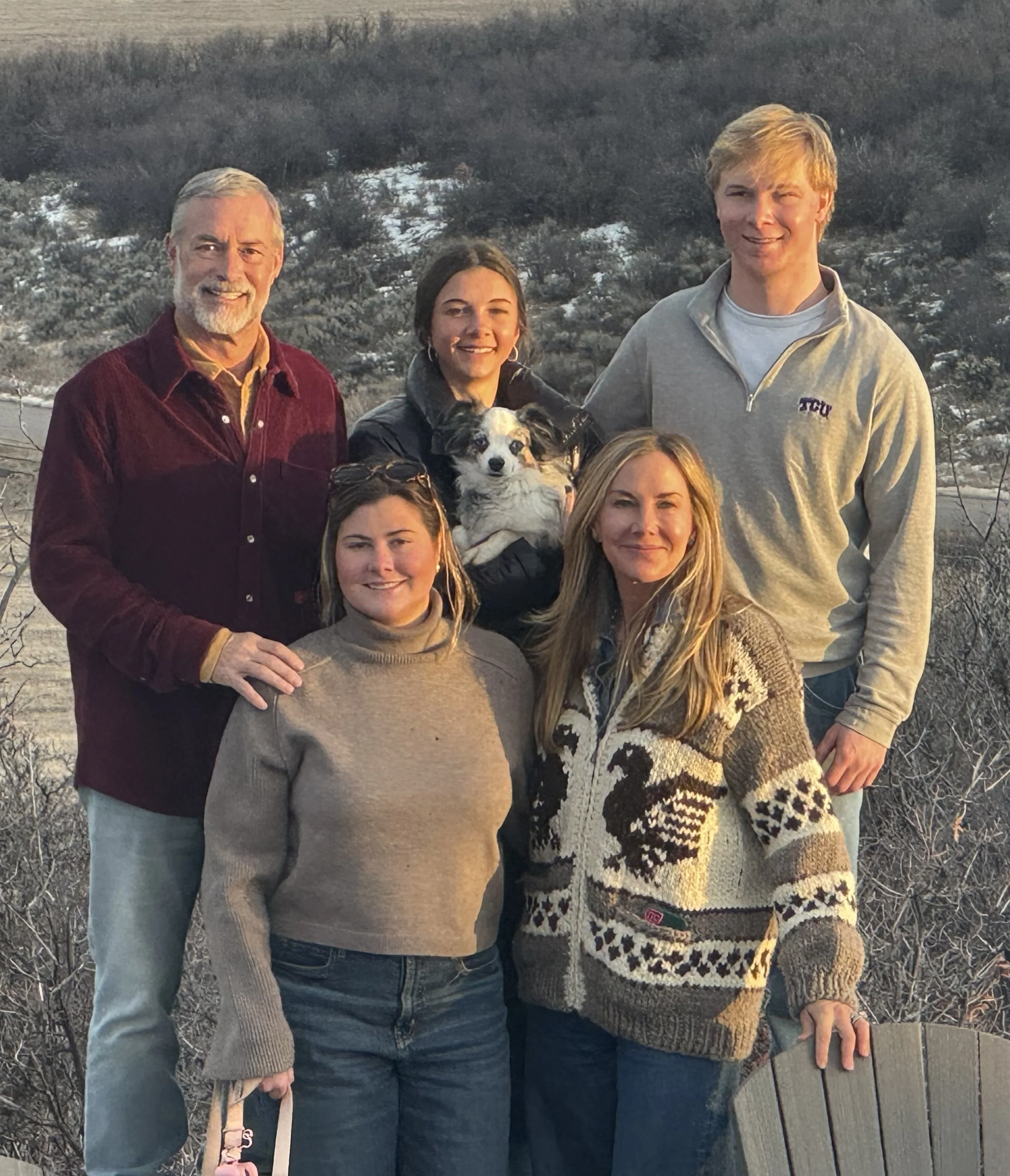 A group of five people and a dog standing outdoors on a winter day with snow-covered trees and hills in the background.