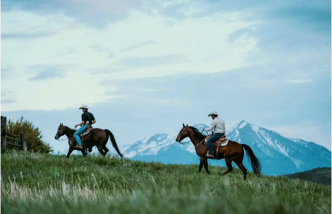 Two people riding horses on a grassy field with snow-capped mountains in the background under a partly cloudy sky.