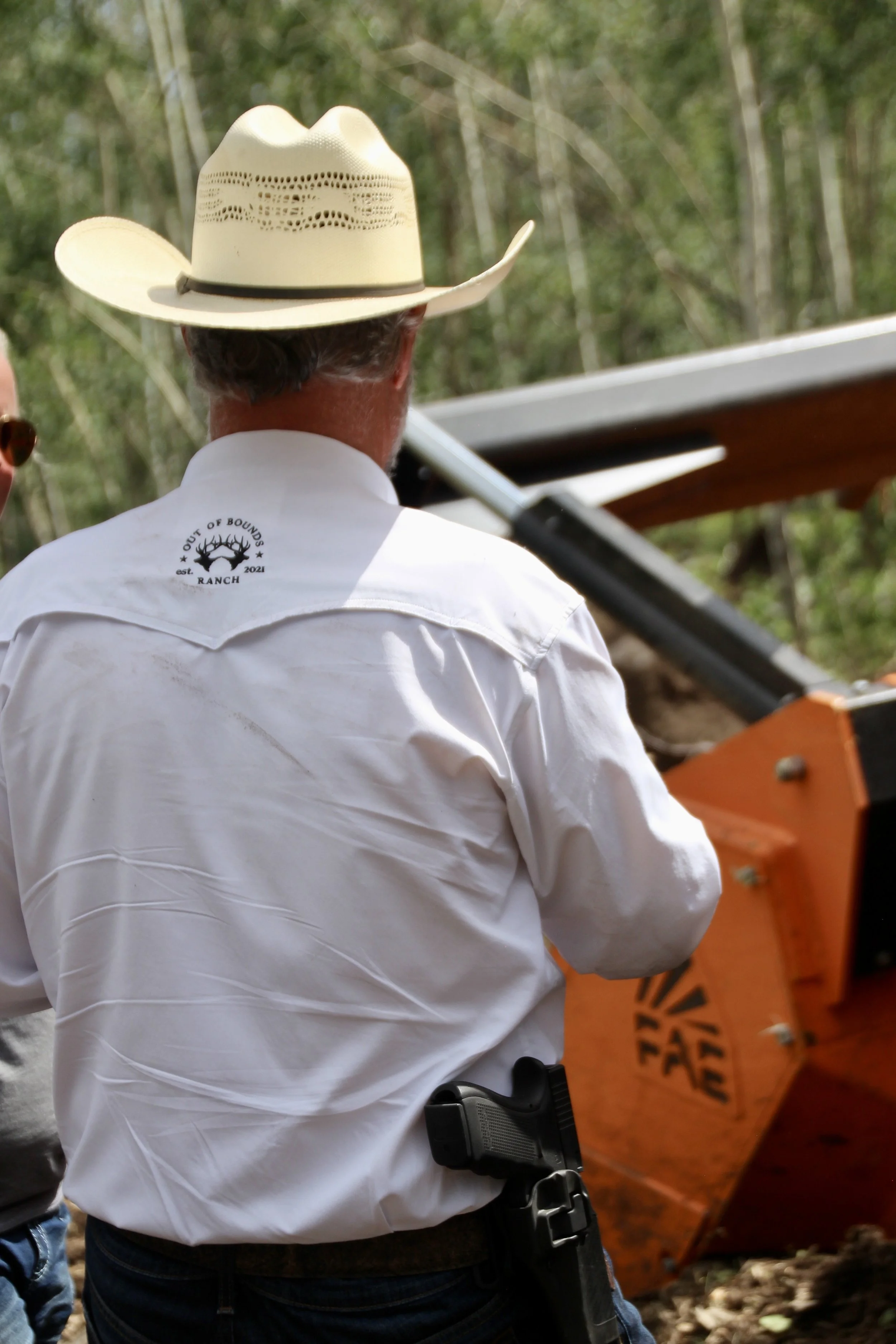 A man wearing a white cowboy hat and a white shirt with a ranch logo on the back is seen from behind, standing outdoors near a wood chipper or similar equipment in a wooded area.