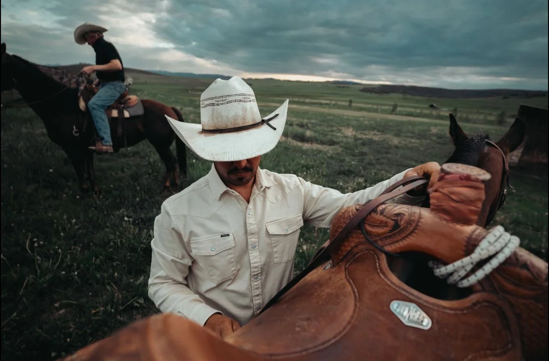 Man wearing a cowboy hat and fringe shirt adjusting a saddle on a horse in an open countryside with another rider on horseback in the background.