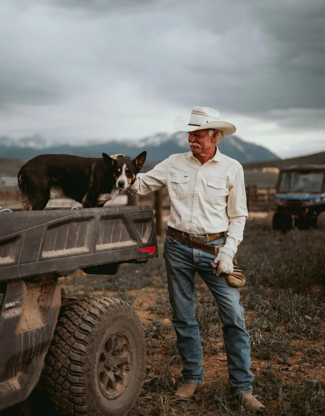 An older man with a cowboy hat and beige long sleeve shirt standing outdoors in a rugged landscape, with mountains in the background. He is petting a dog on the back of a black truck.