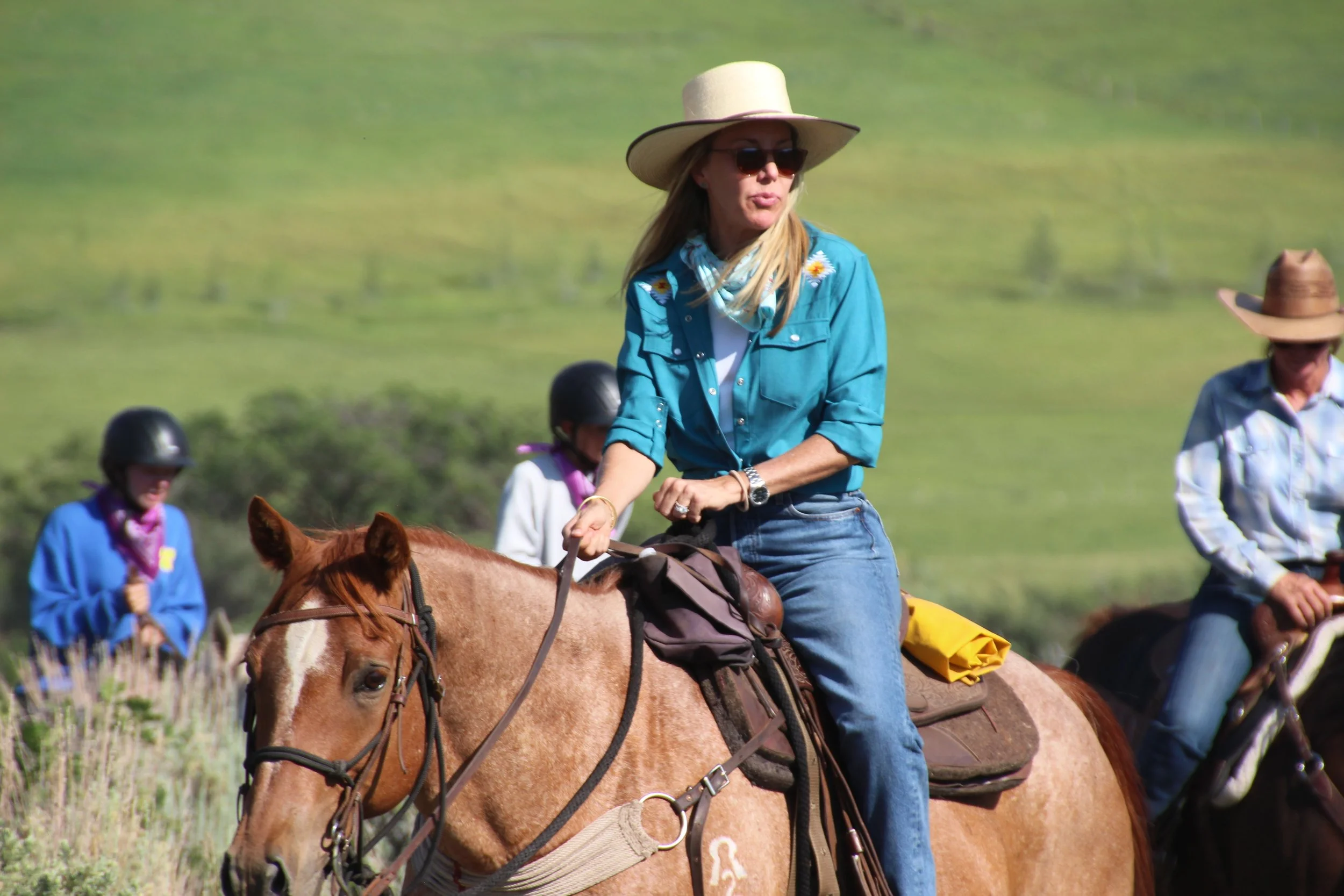 A woman riding a horse outdoors with a green hillside background, wearing a blue shirt, jeans, sunglasses, and a wide-brimmed hat. Other people on horses are visible in the background.