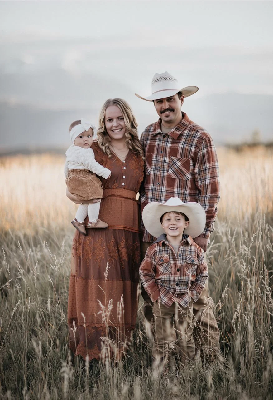 A family of four dressed in western wear standing in a field of tall grass with a cloudy sky in the background.