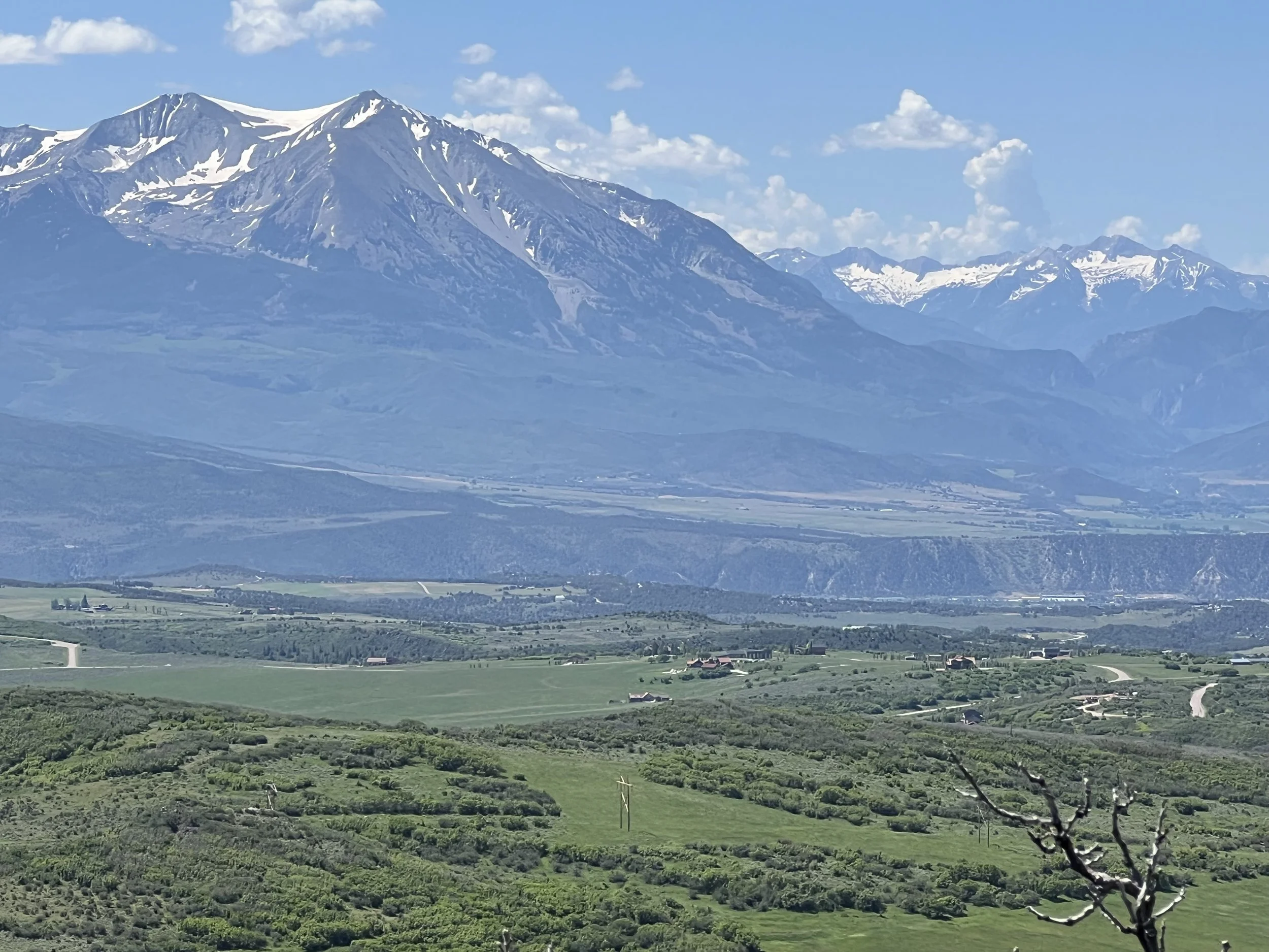 Scenic view of snow-capped mountains in the background, with green fields and scattered houses in the foreground under a partly cloudy sky.