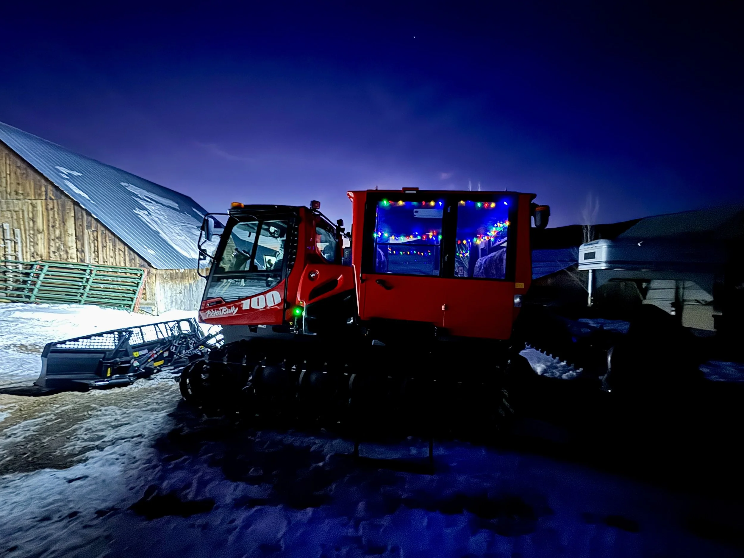 A red snow grooming machine decorated with colorful Christmas lights parked on a snowy surface during dusk or night, with a rustic barn and another trailer in the background.