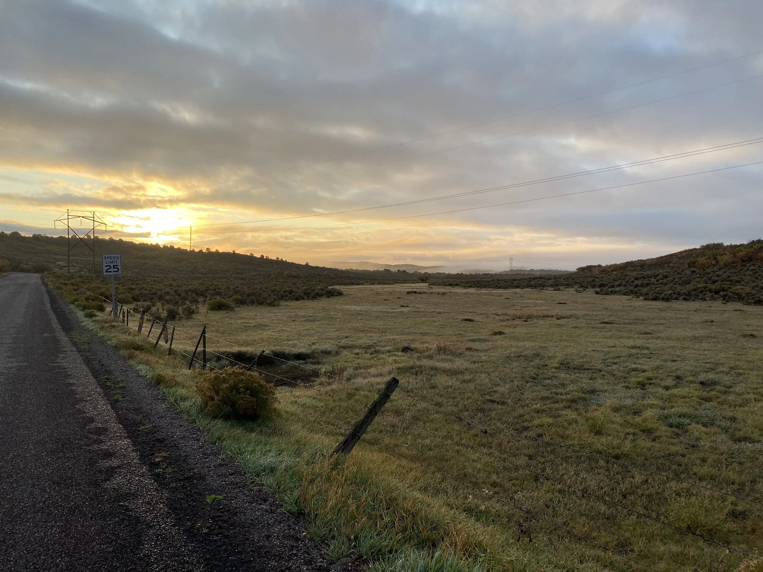 A rural landscape at sunset showing a paved road on the left, a fence, grassy fields, hills, and power lines under a cloudy sky with the sun near the horizon.
