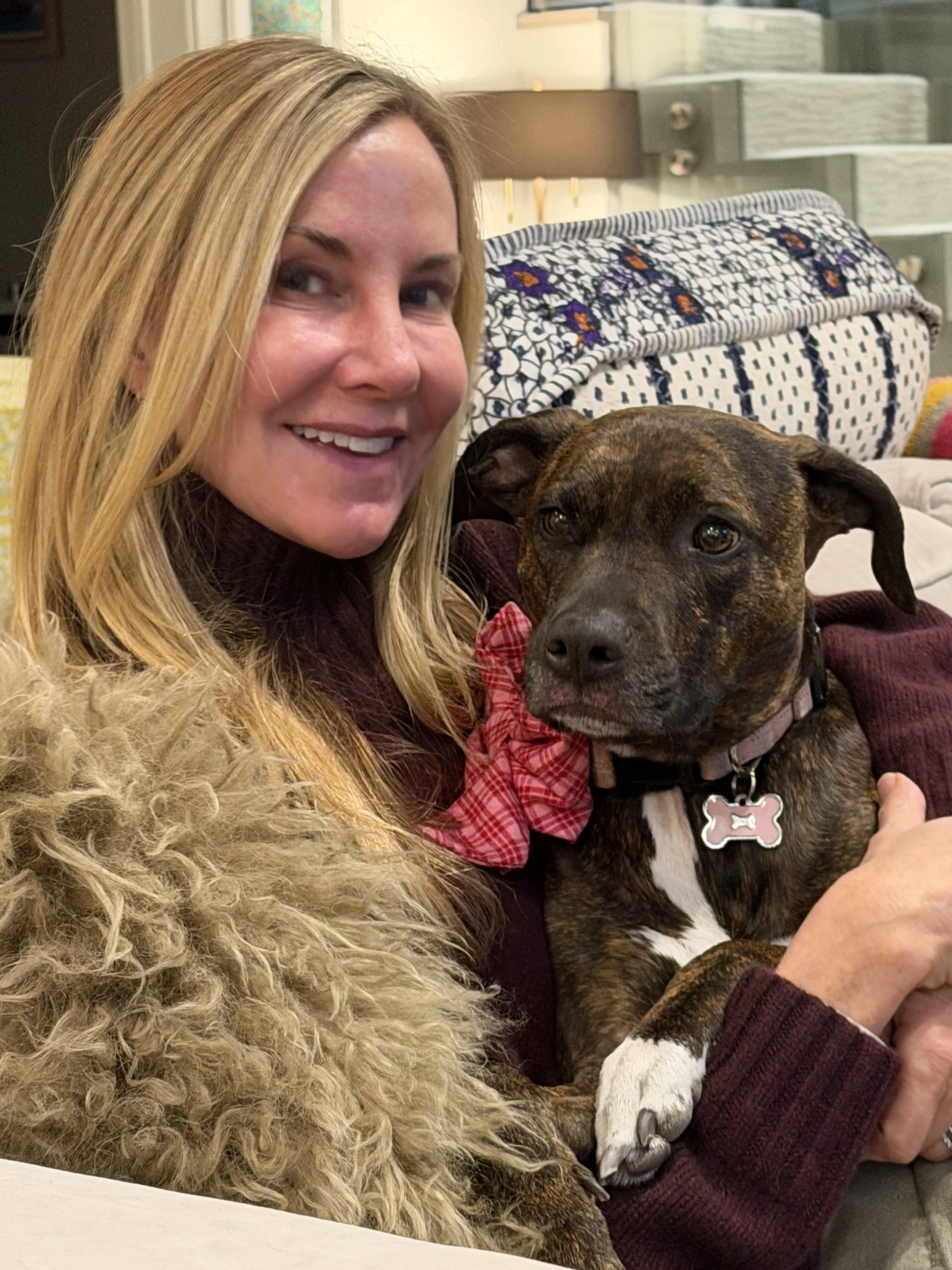 A woman with blonde hair smiling, sitting on a sofa with a brindle-coated dog wearing a pink collar and bow, and holding its paw. The background shows shelves and a lamp.