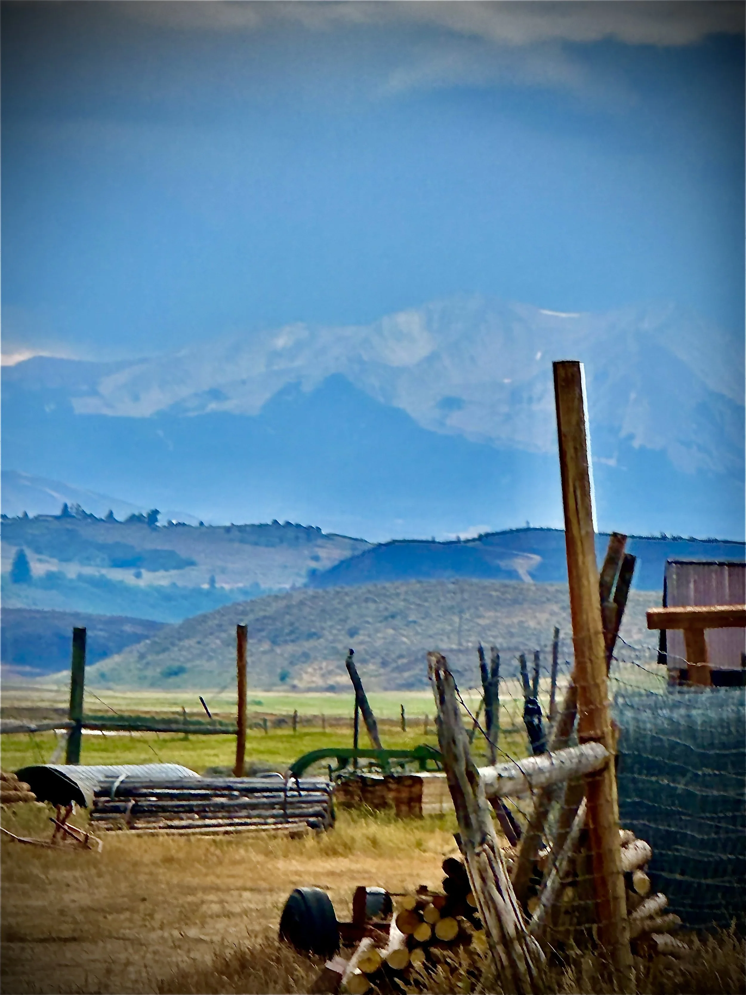 A rural landscape with mountains in the background, a farm with logs and fencing in the foreground, and a blue sky overhead.
