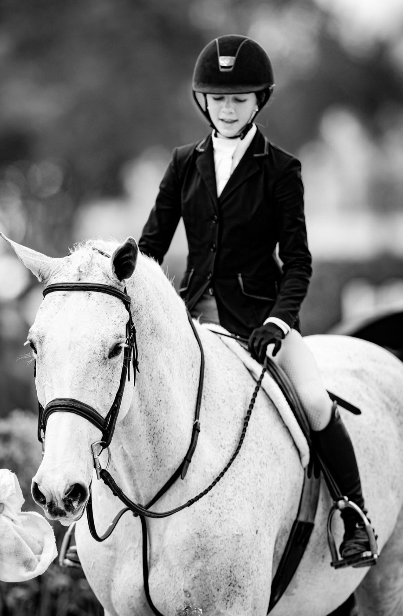 A young equestrian woman in riding attire, including a helmet and jacket, riding a white horse.