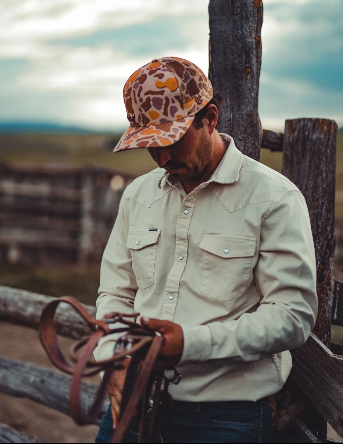 A man leaning against a wooden fence, looking down at a pair of sunglasses, wearing a camouflage cap and a light beige button-up shirt.