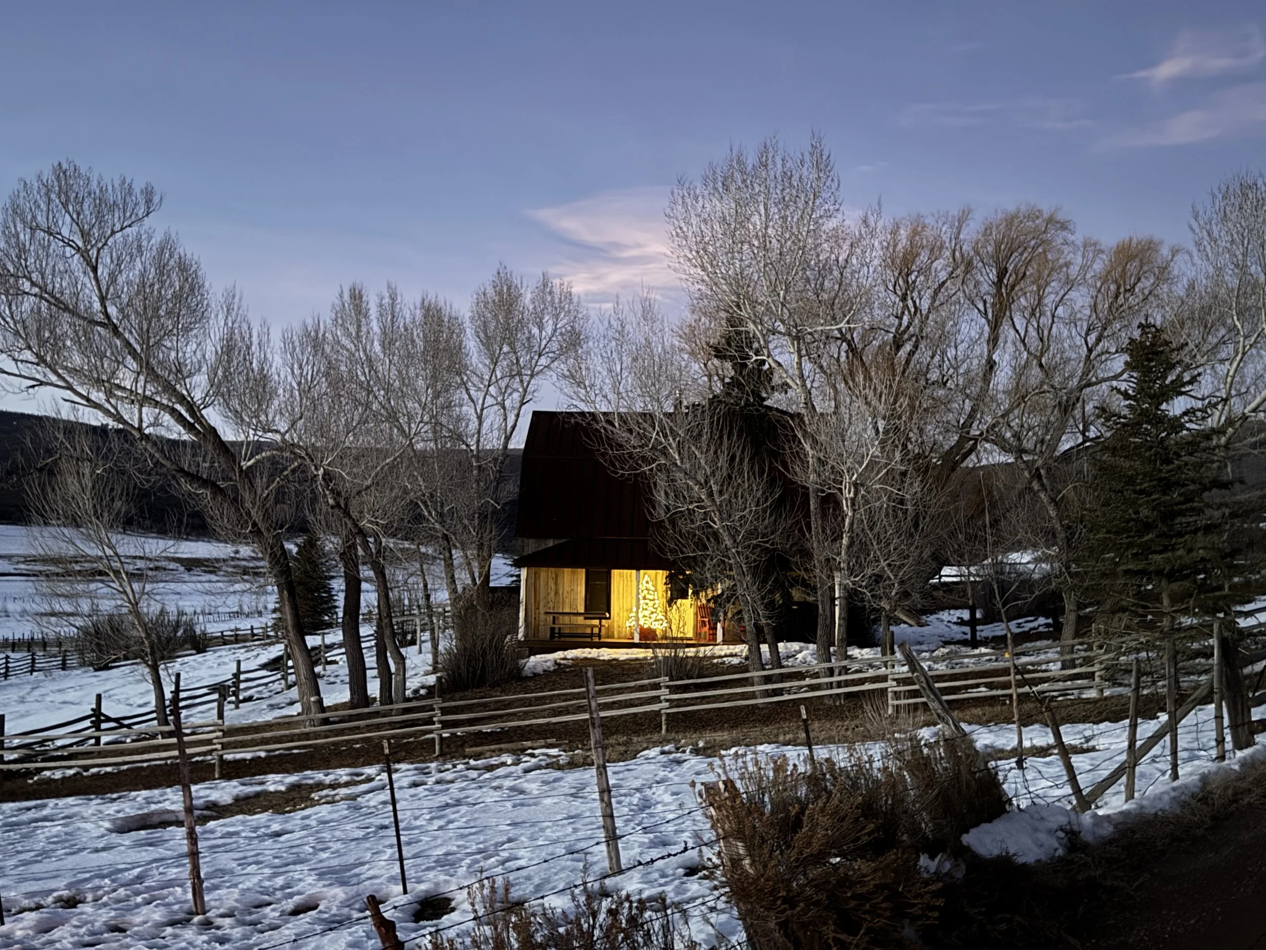 A winter scene with snow on the ground, leafless trees, a rustic wooden house with warm lighting, and a faintly visible Christmas tree decoration near the entrance, set against a backdrop of a blue sky with some clouds.