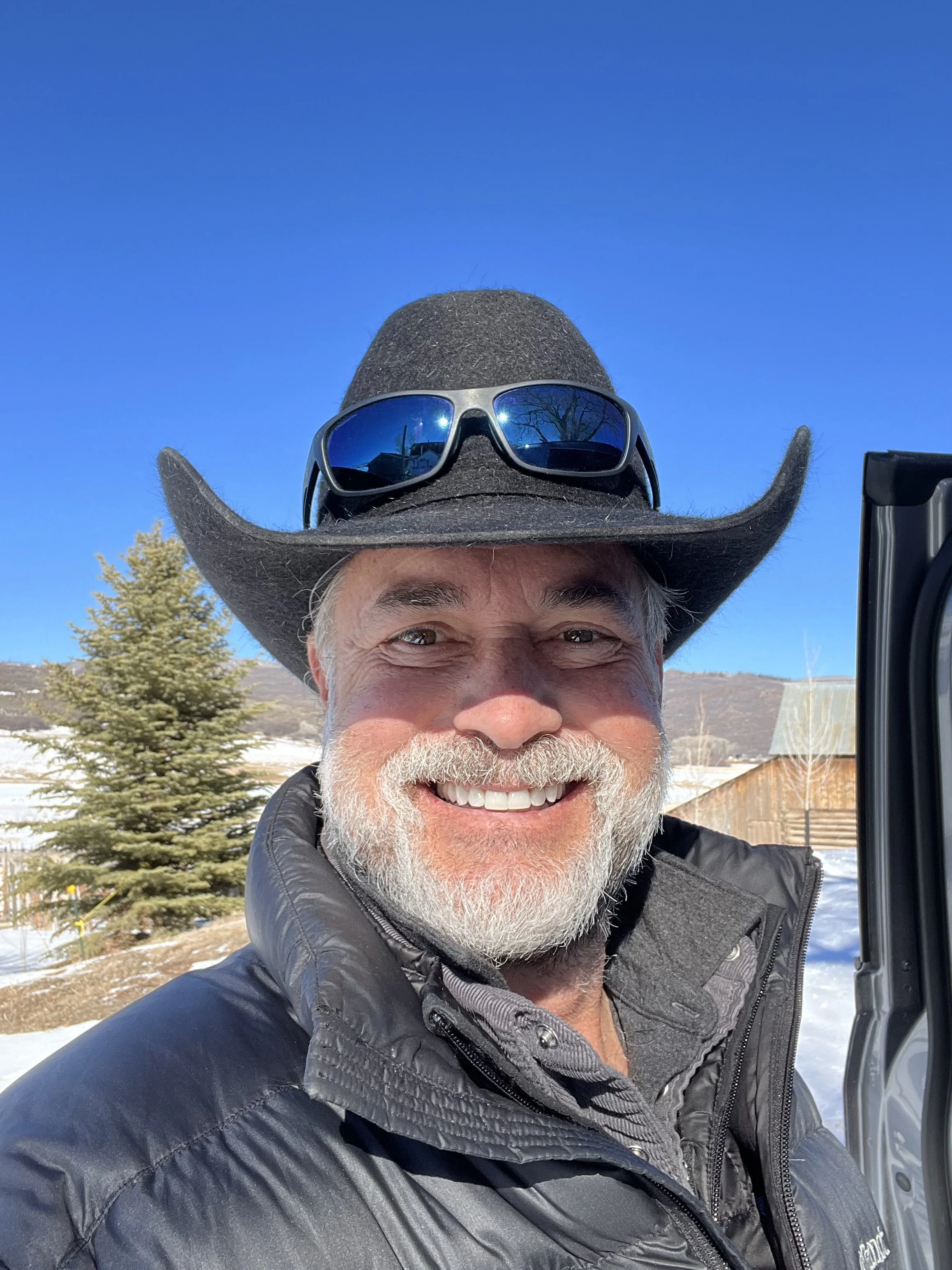 A smiling man with a gray beard wearing a black cowboy hat and sunglasses, standing outdoors in a snowy landscape, with trees and a building in the background under a clear blue sky.