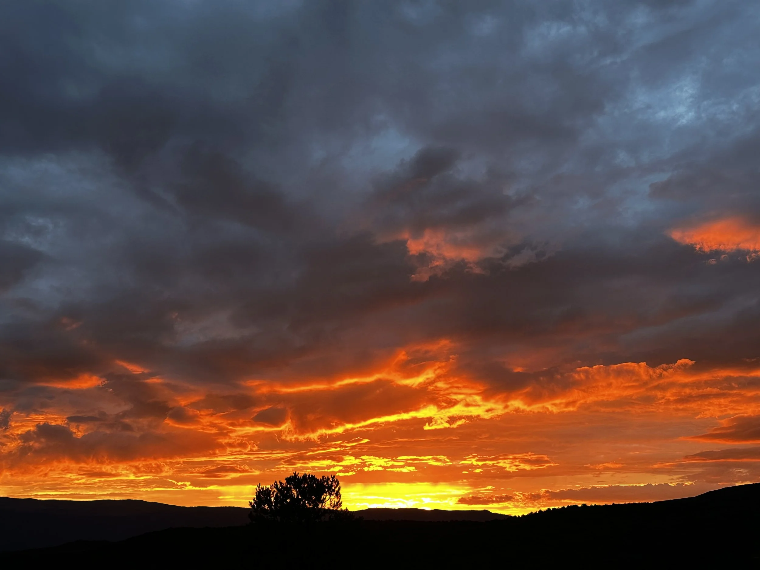 Colorful sunset sky with dark clouds, orange and yellow hues, silhouette of trees and hills.