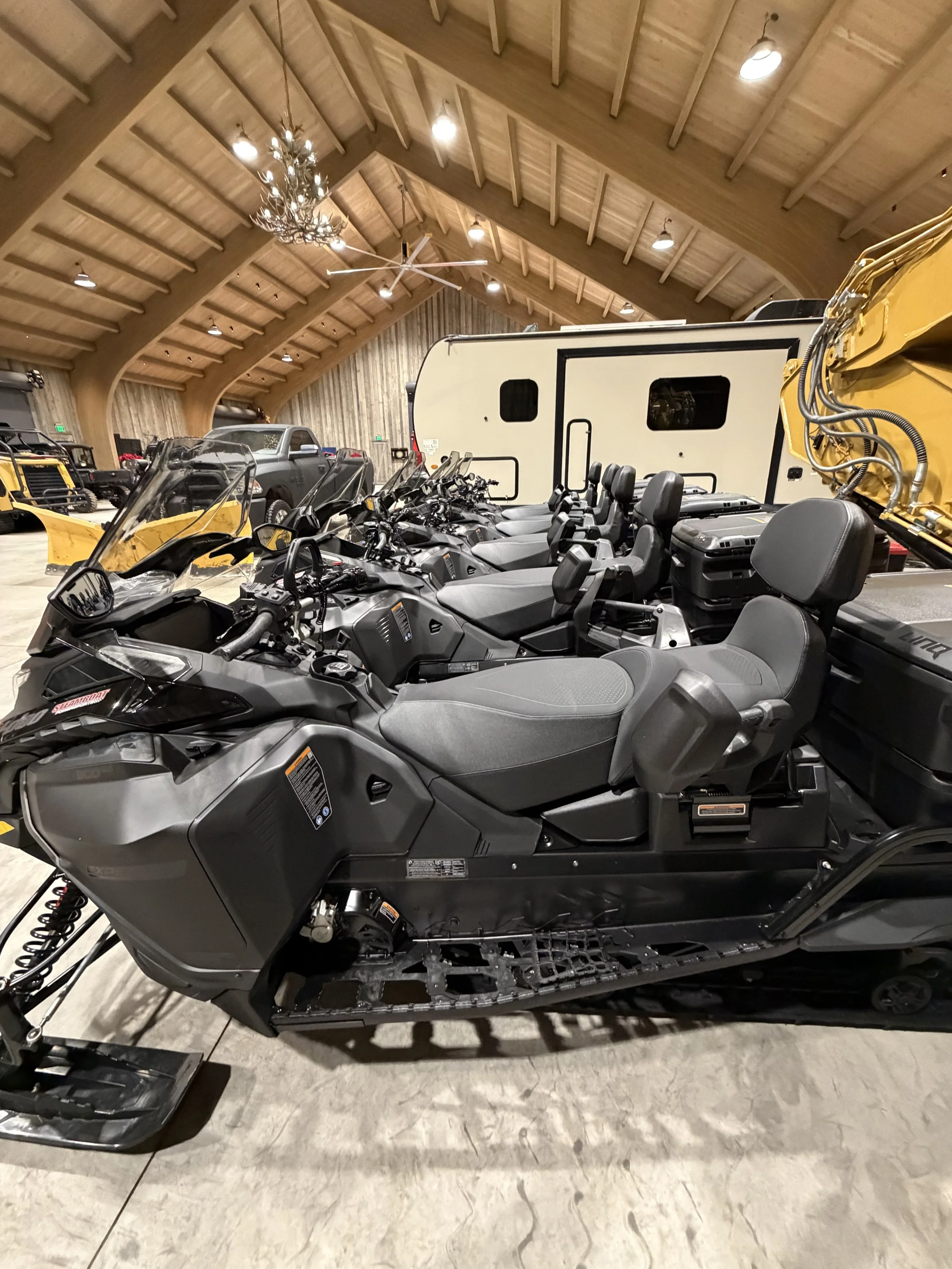 Indoor showroom with several snowmobiles parked in a row, surrounded by recreational and utility vehicles, under a wooden vaulted ceiling with pendant and chandelier lighting.