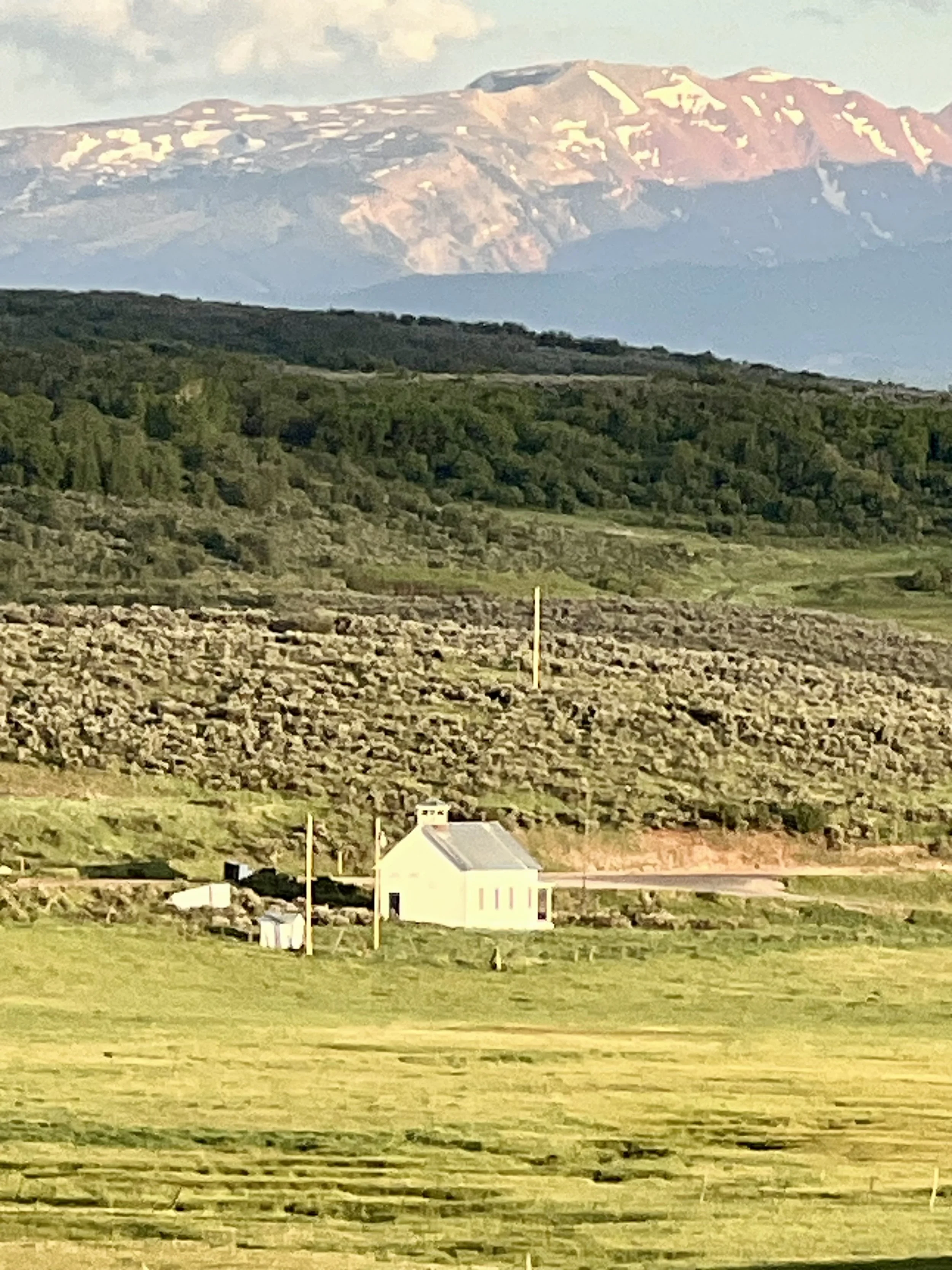 A small white building with solar panels on the roof, surrounded by green fields, with rolling hills and a snow-capped mountain in the background.