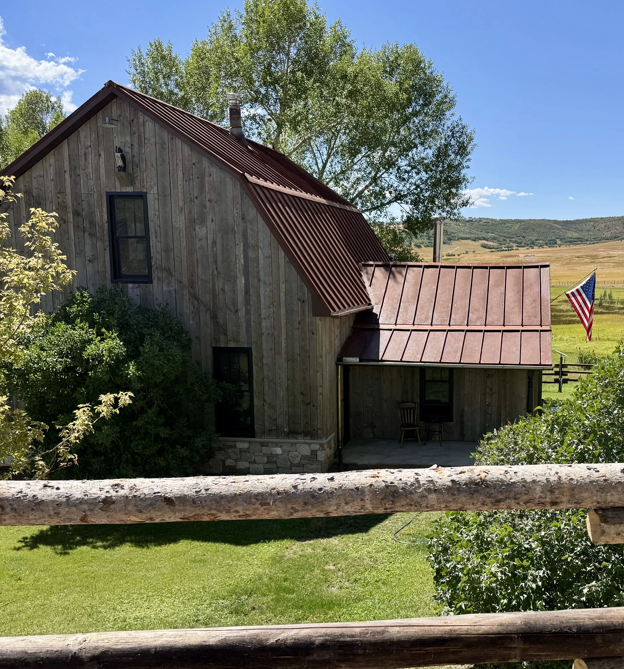A rustic wooden house with a red metal roof, surrounded by green trees and shrubs, with a wooden balcony and an American flag flying in the background under a blue sky.