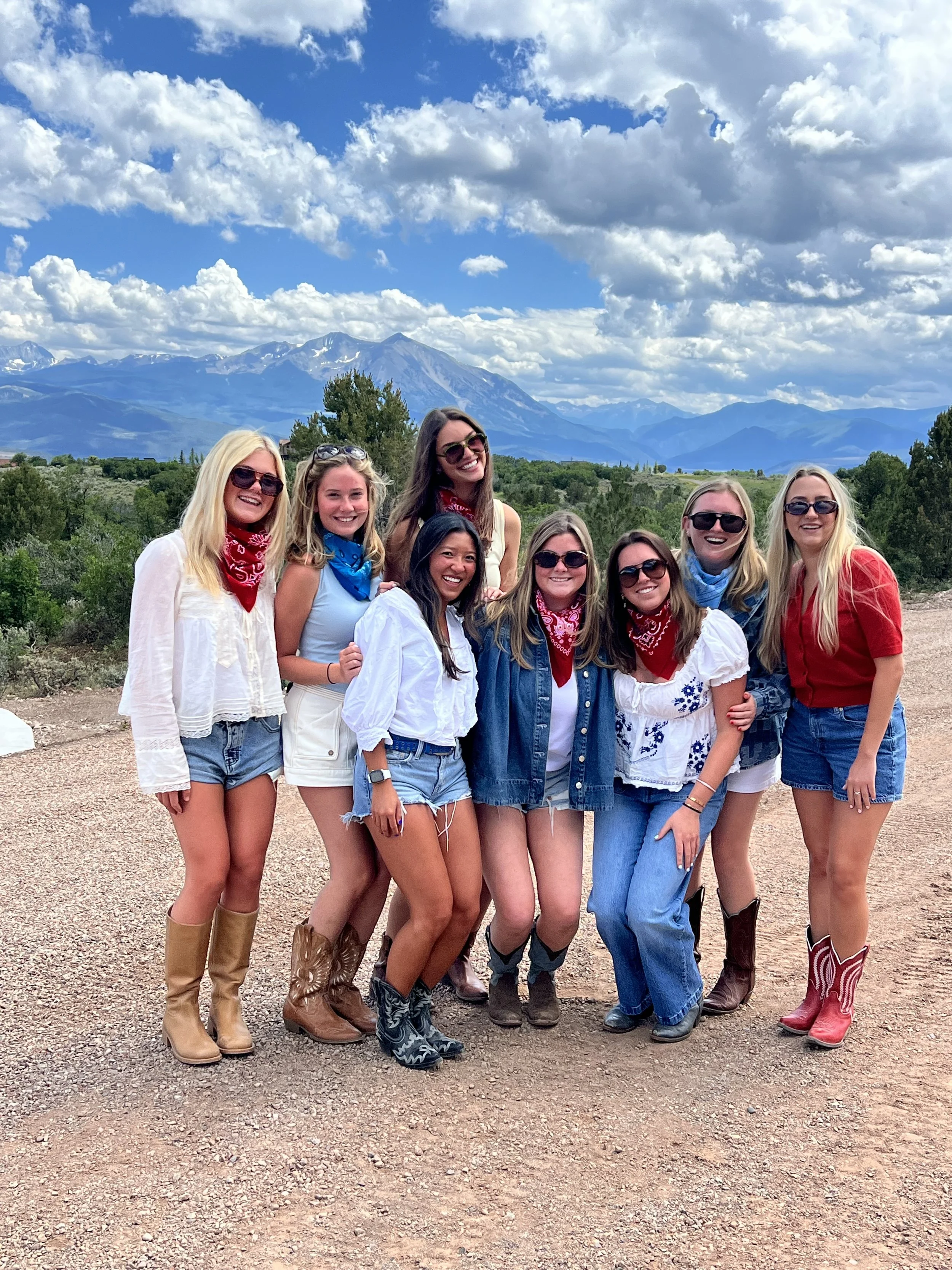 Group of eight women dressed in casual country-style clothing, wearing bandanas, jeans, and cowboy boots, standing outdoors on a dirt path with mountains and sky in the background.