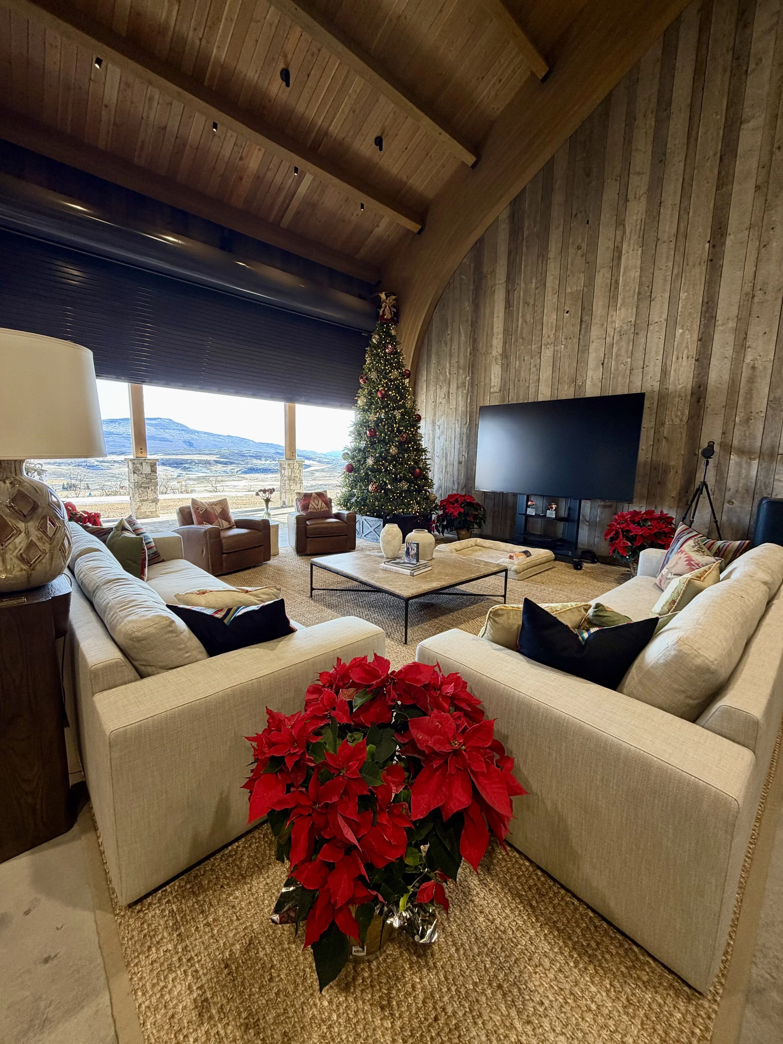A cozy living room decorated for Christmas with a large Christmas tree, red poinsettias, and festive pillows on cream-colored sofas. There are armchairs by a large window showing a mountain landscape.