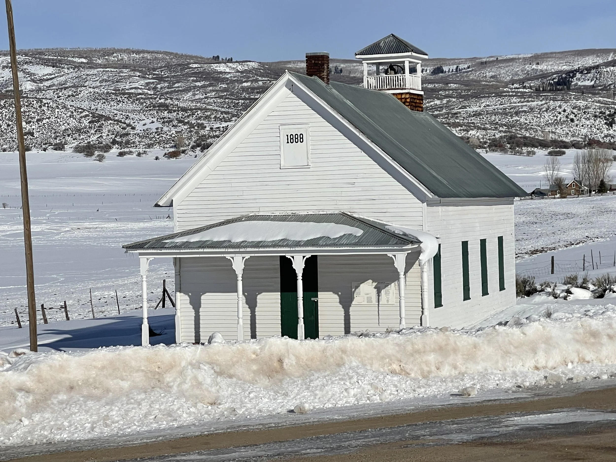 A white wooden historic school house with a porch and a secretary window marked 1888, snow-covered landscape, mountains in the background, and a small building near the house.  Carbondale history.