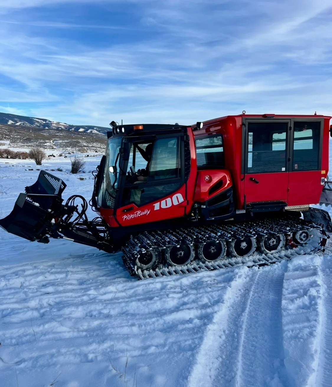 Red snowcat vehicle with tracks on snow-covered terrain, mountains in the background, and a cloudy blue sky.