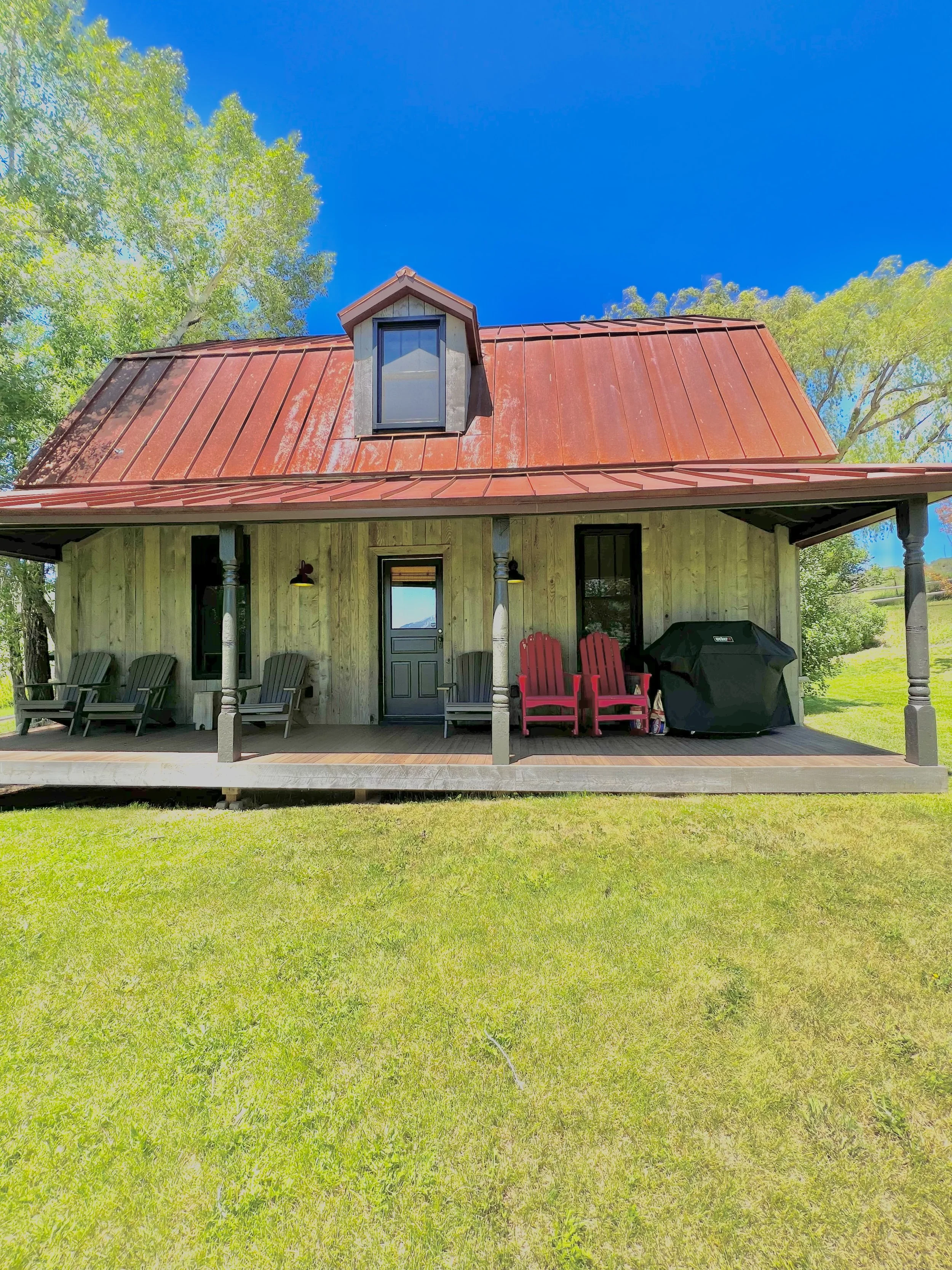 A house with a red metal roof, weathered wooden siding, and a front porch with chairs and a barbecue grill, set against a blue sky and green trees.
