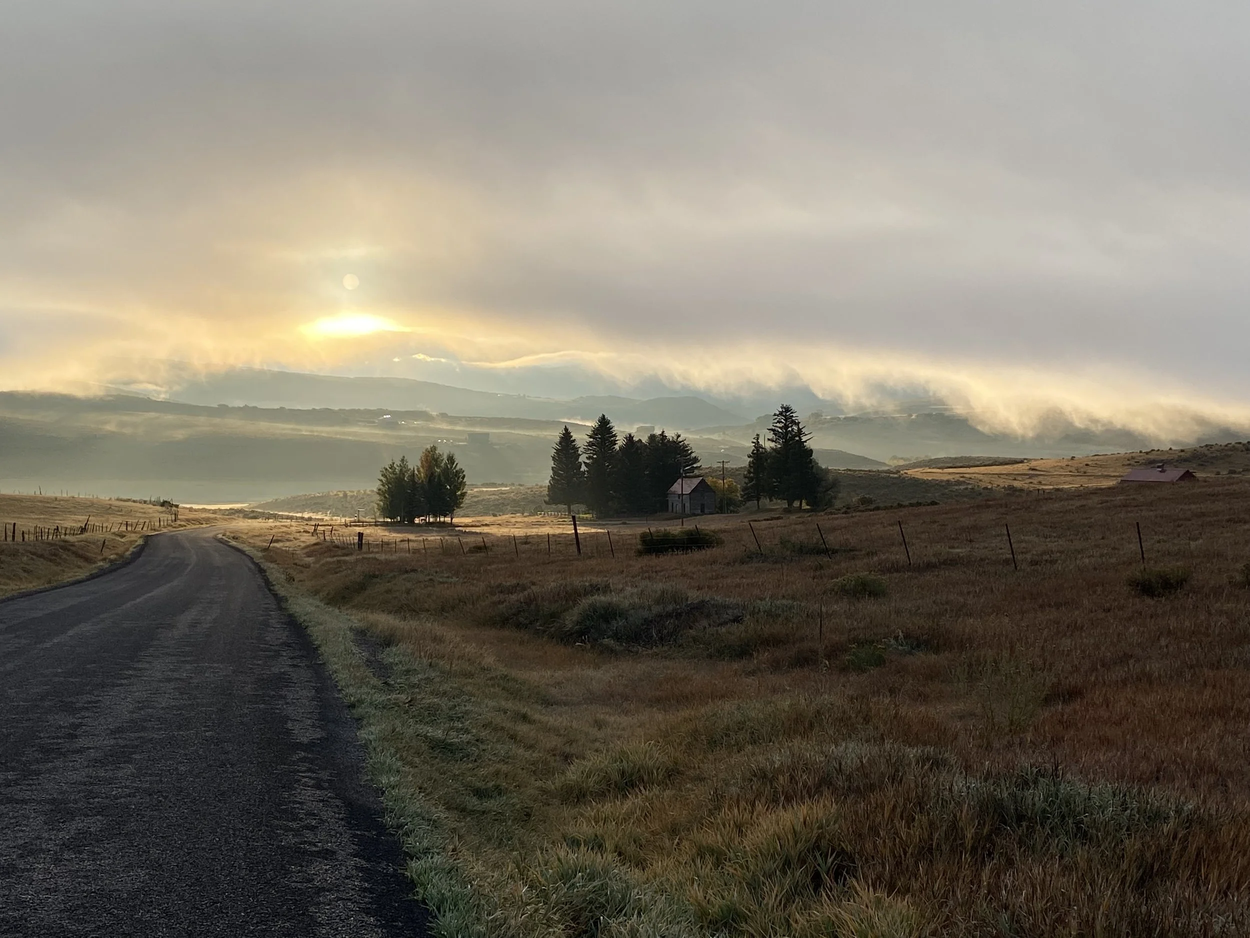A winding rural road runs through a grassy field with a few trees and small farm buildings. Cloudy sky with sunlight peeking through, casting a warm glow over the landscape.